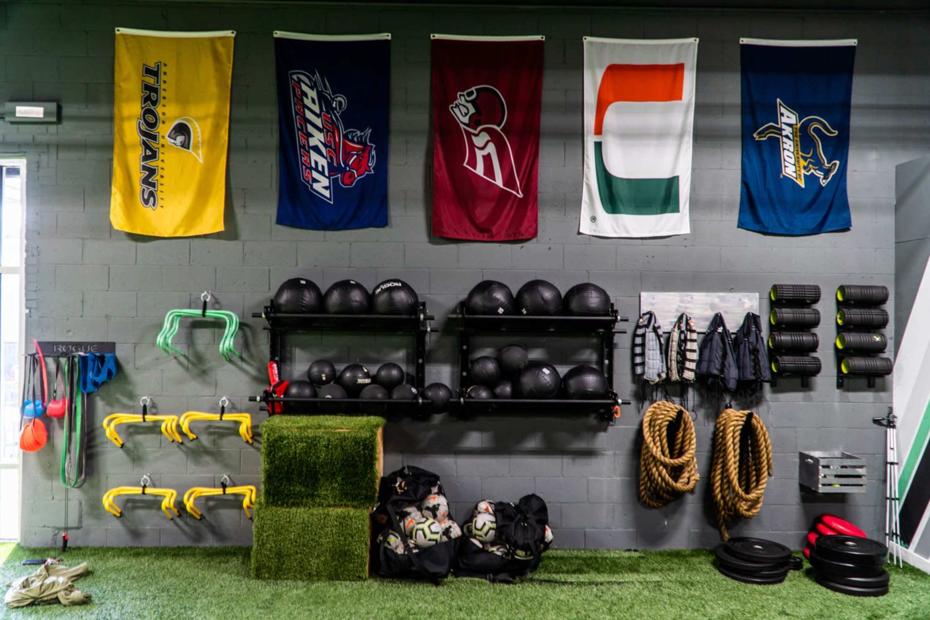 A gym wall adorned with various team flags, with exercise equipment including medicine balls, ropes, and training gear organized on shelves and hooks.