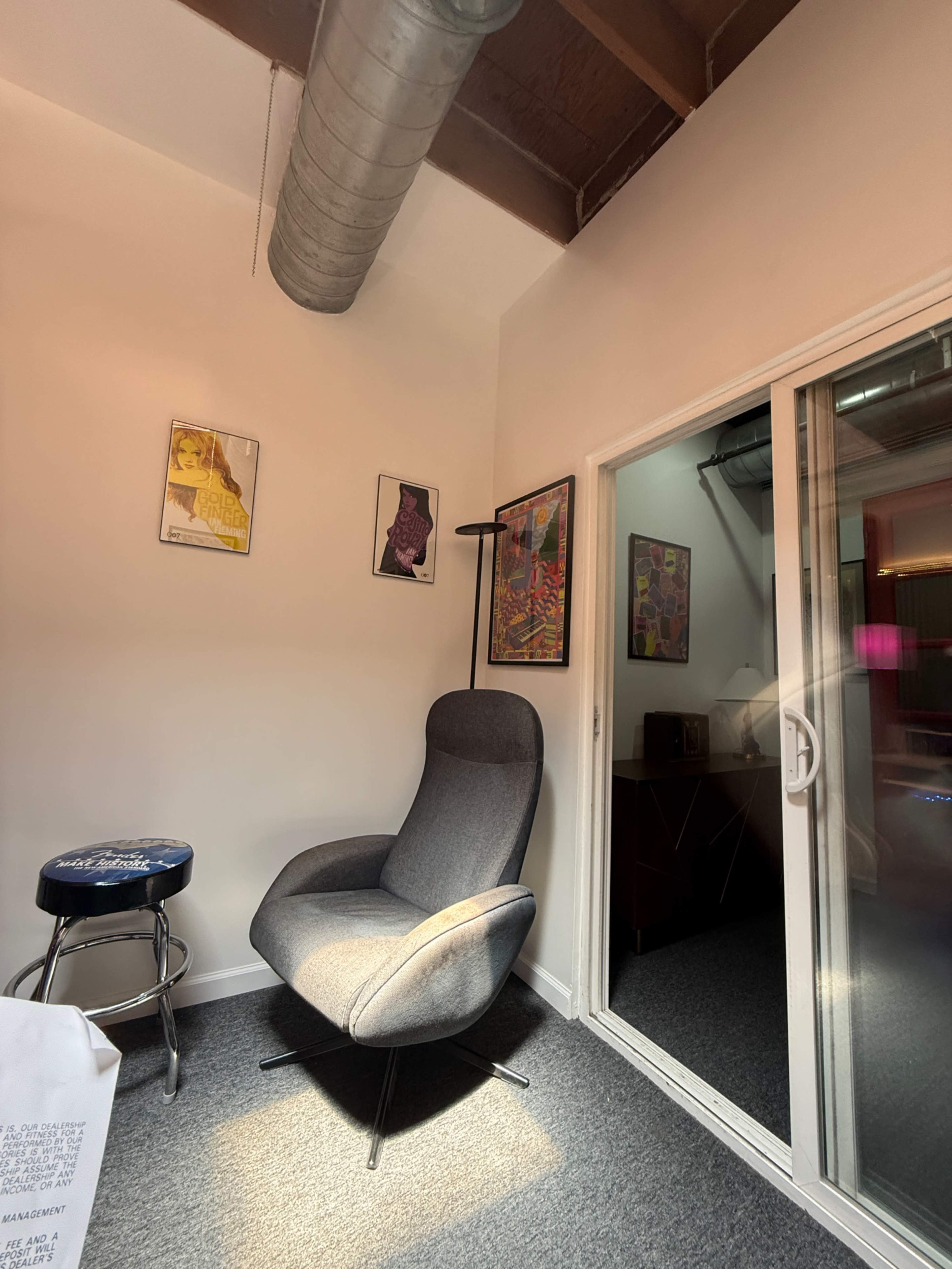 The image shows a small room with a gray chair, a stool, and artwork on the walls, featuring a sliding glass door on one side.
