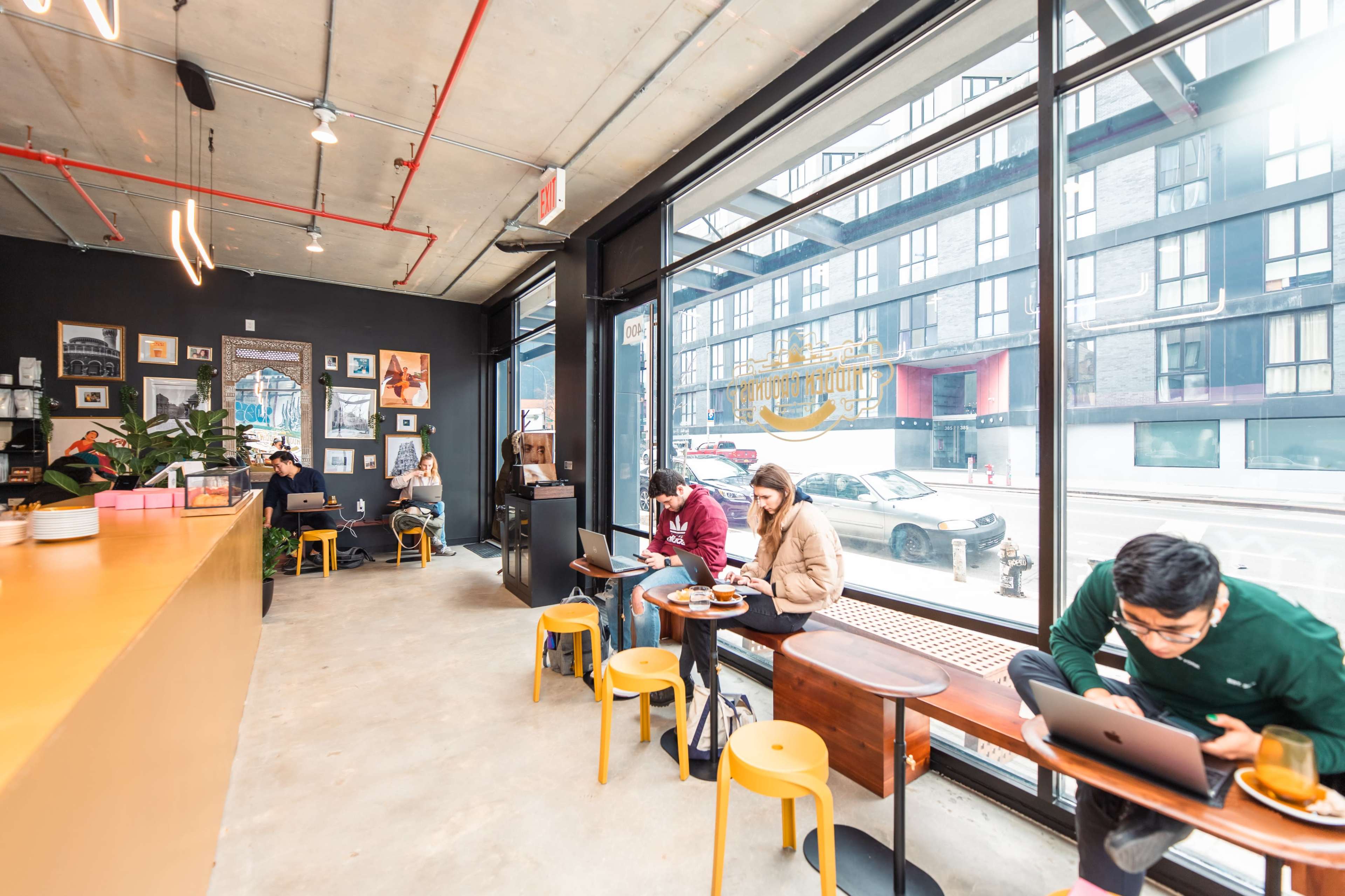A cozy café interior features patrons seated at tables, some working on laptops, while large windows offer a view of the street outside.