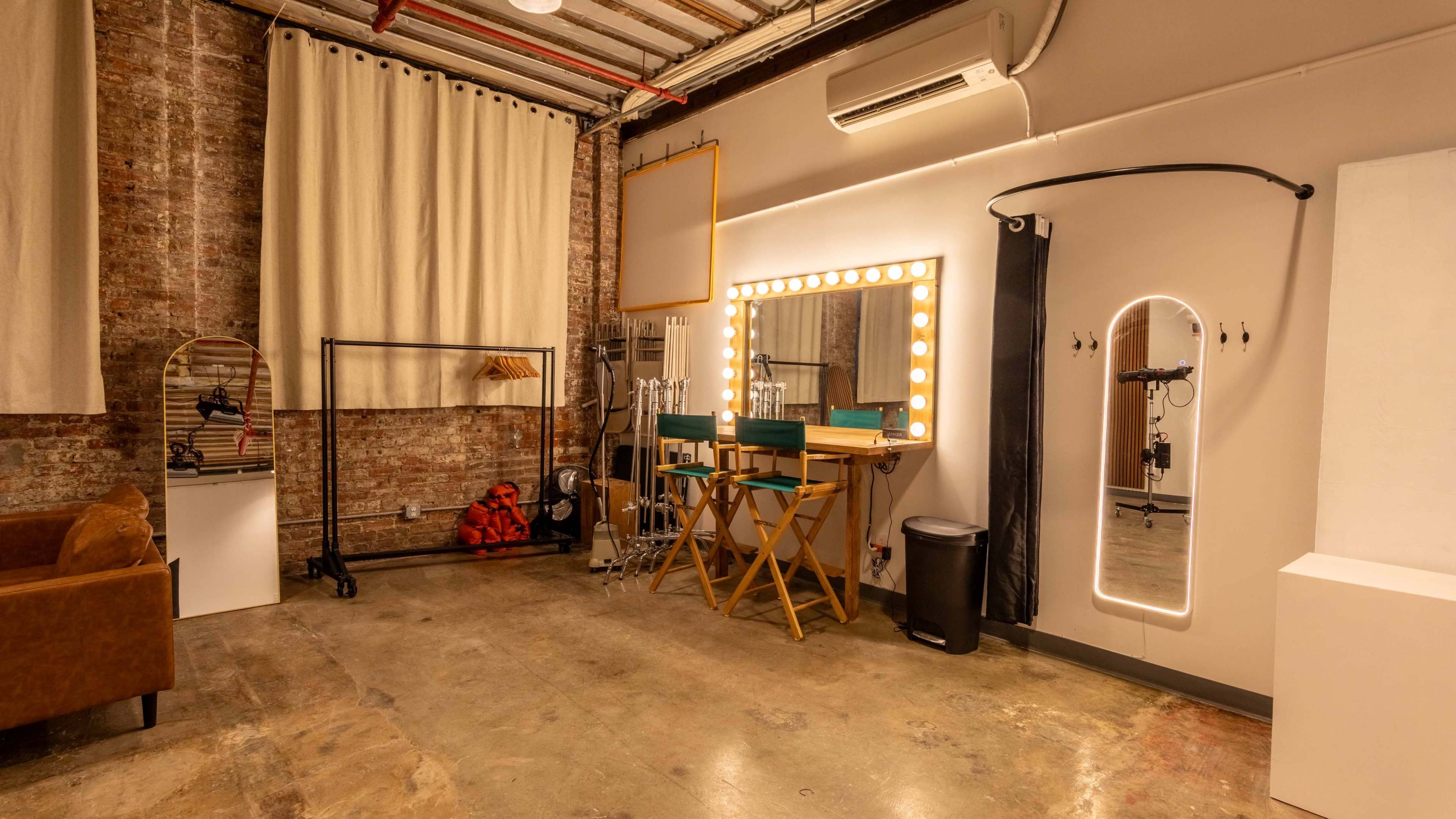 The image shows a dressing room with a large illuminated mirror, two director-style chairs, and various styling equipment against a brick wall.