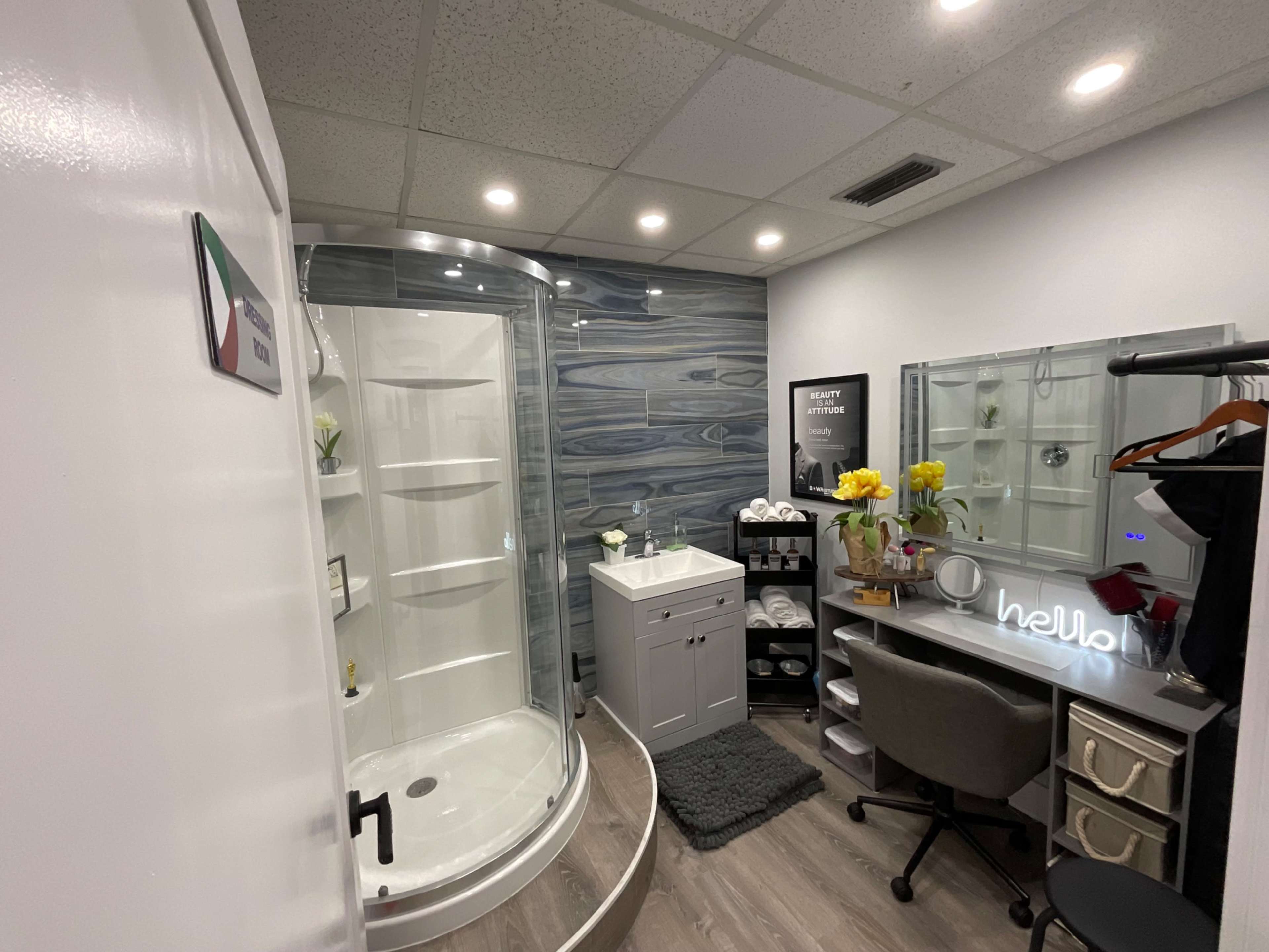 A modern bathroom with a curved shower, a white vanity with towels, a seating area with a mirror and a "hello" light sign.