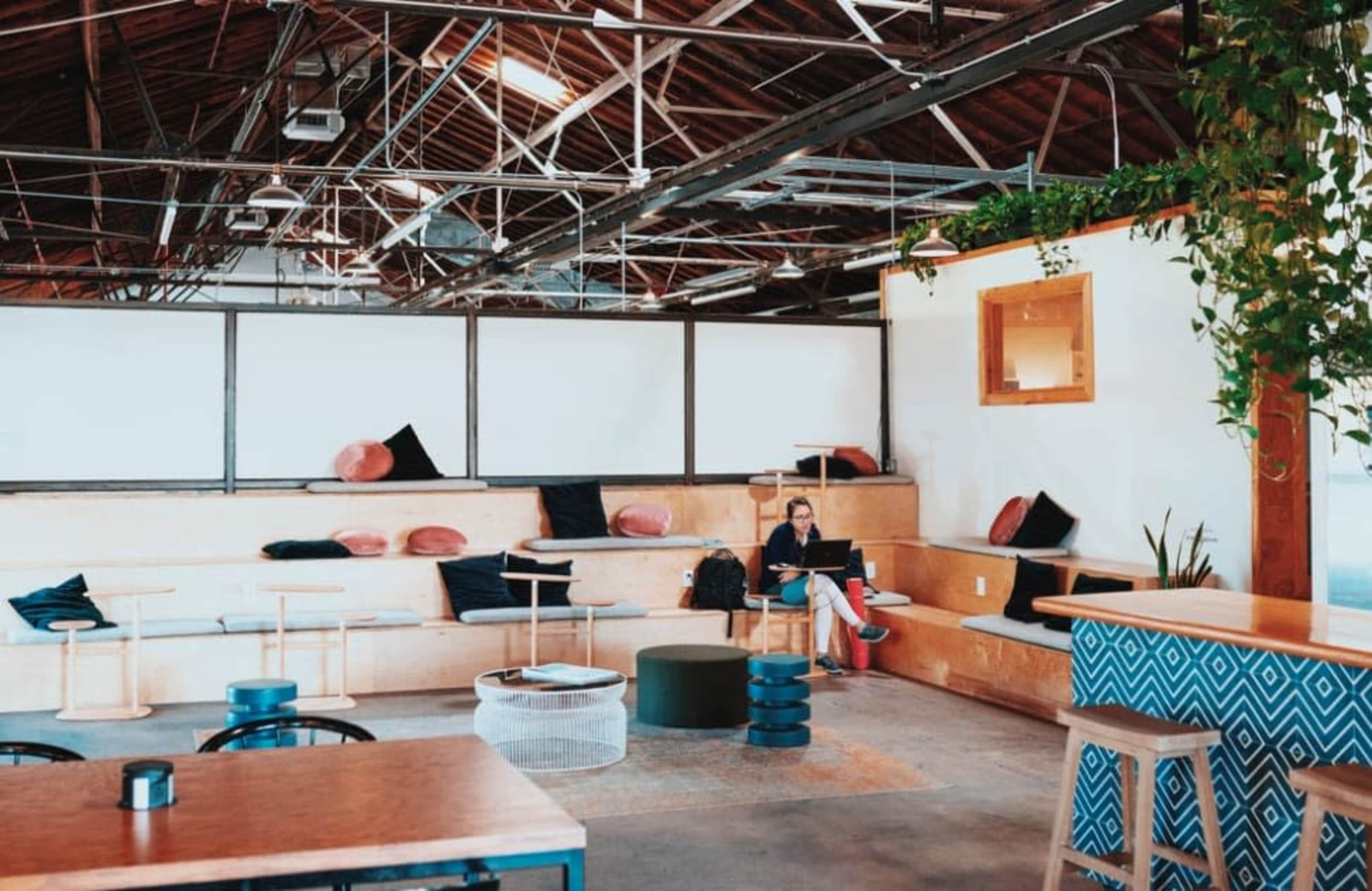 A person is sitting on a wooden bench in a large, open space with high ceilings, working on a laptop among various seating arrangements.