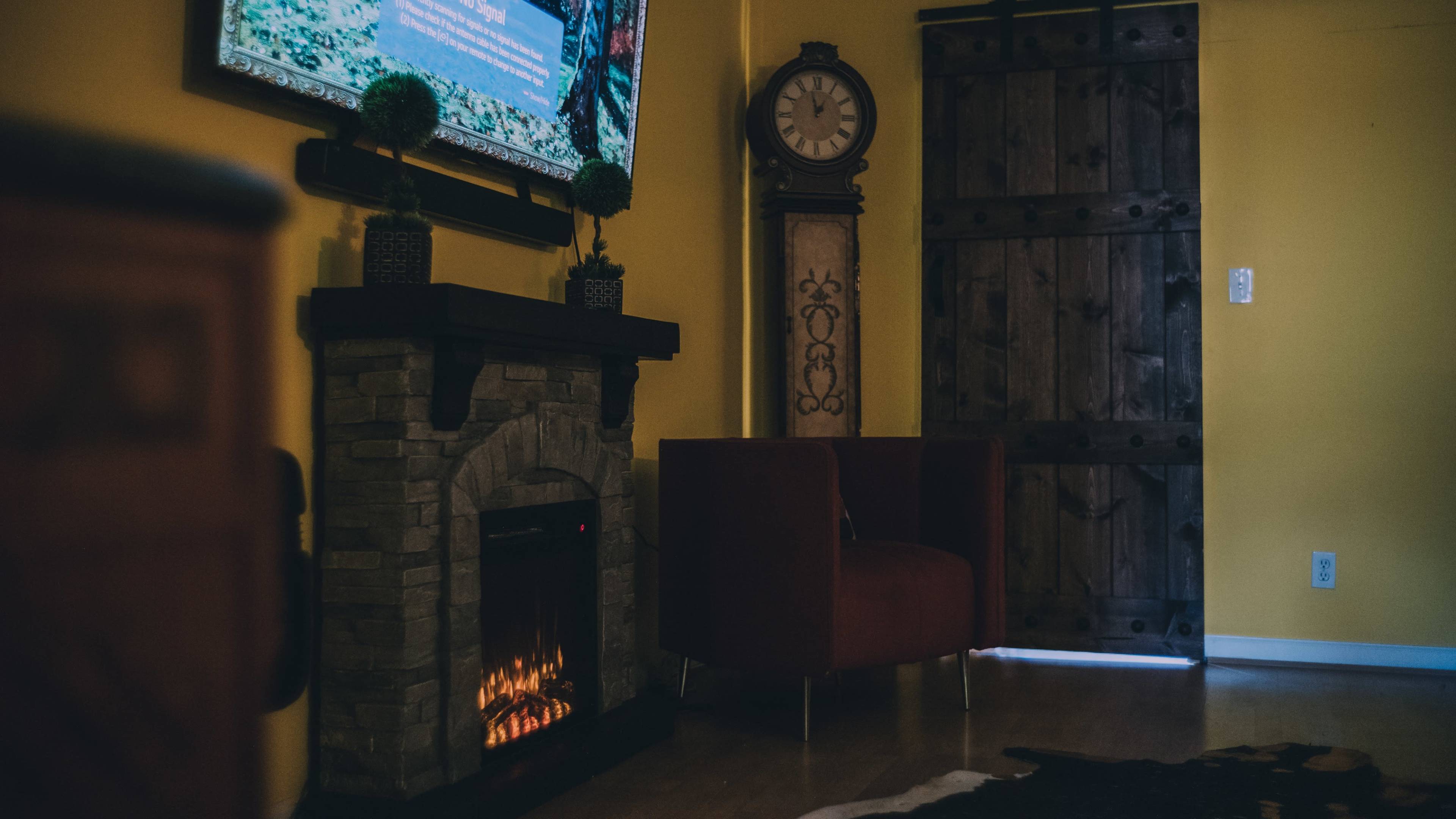 The image shows a cozy living room with a stone fireplace, a red armchair, a large wall clock, and a wooden door.