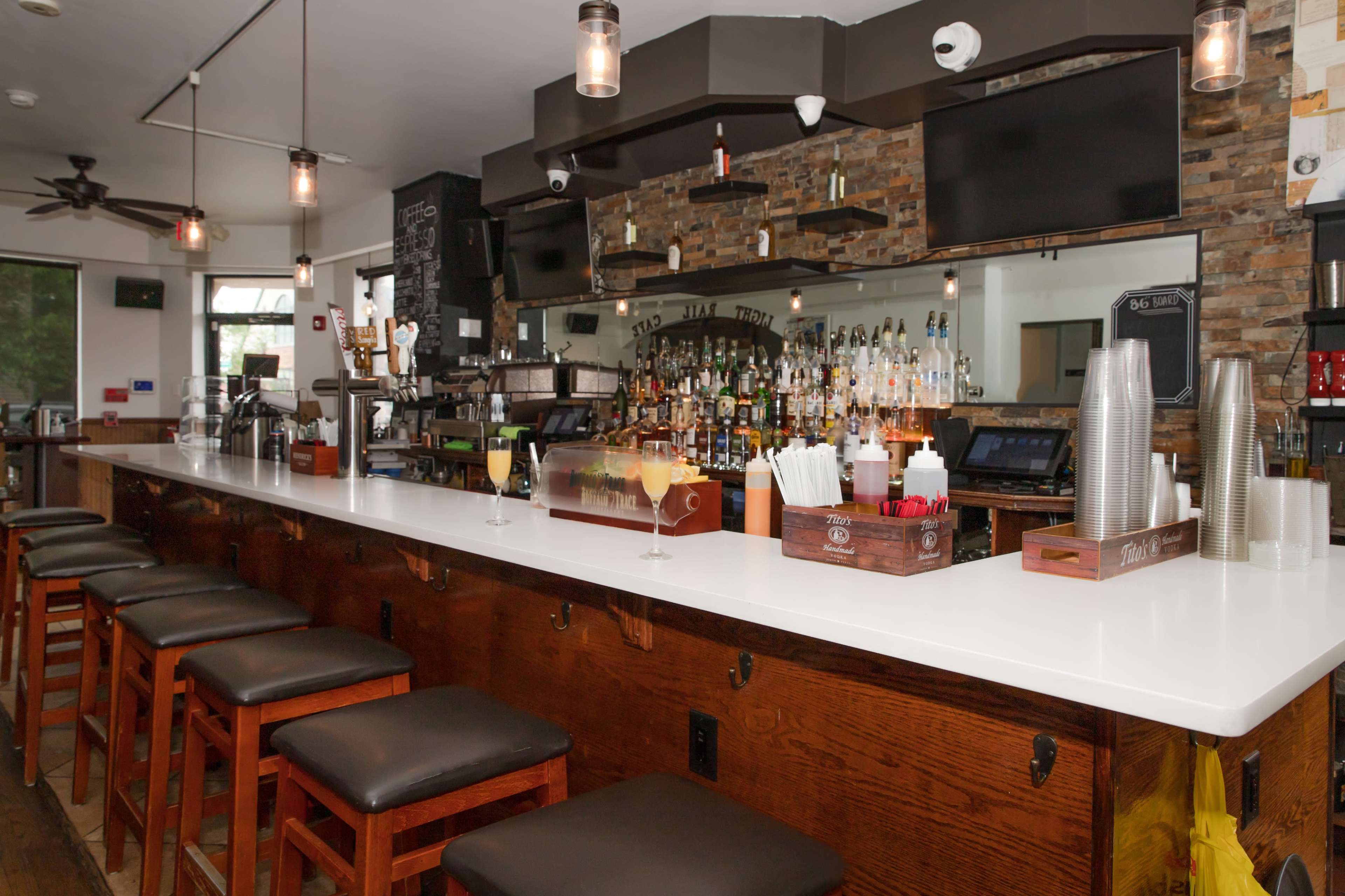 A well-lit bar with a sleek white countertop, wooden stools, and a variety of spirits displayed on shelves behind the counter.