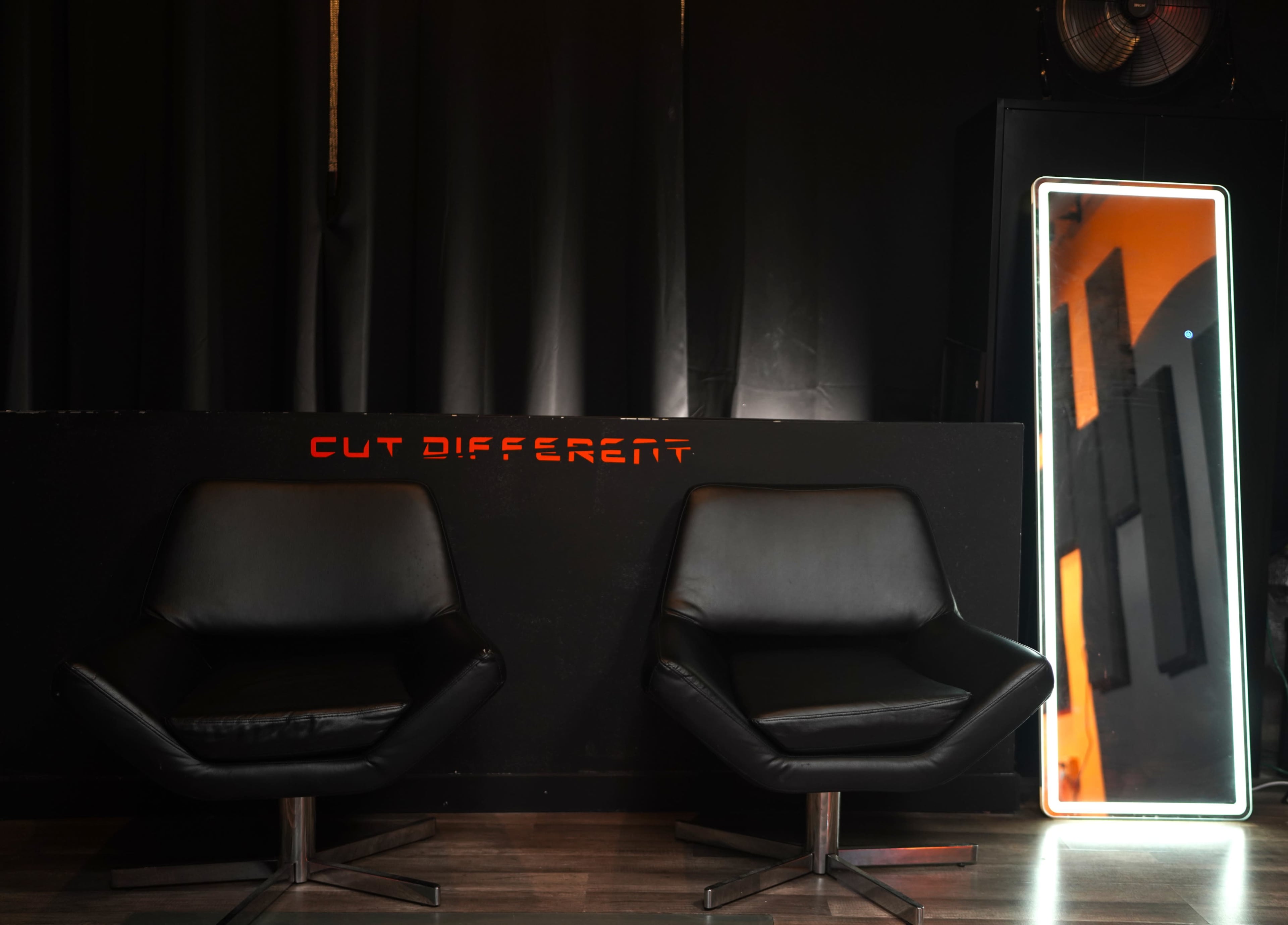The image shows two black chairs positioned in front of a sleek, modern wall featuring the words "CUT DIFFERENT" in illuminated letters, alongside a tall illuminated mirror reflecting the backdrop.