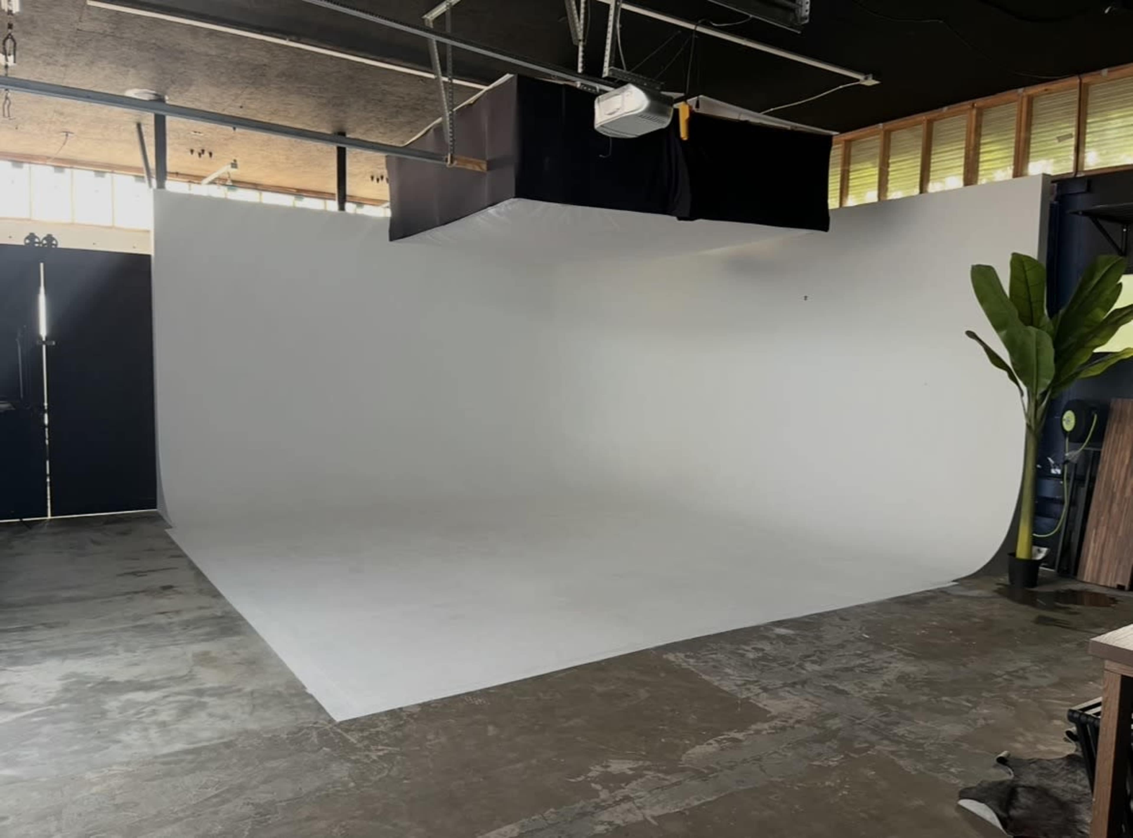 The image shows a spacious photography studio with a white backdrop, polished concrete floor, and a large window letting in natural light.