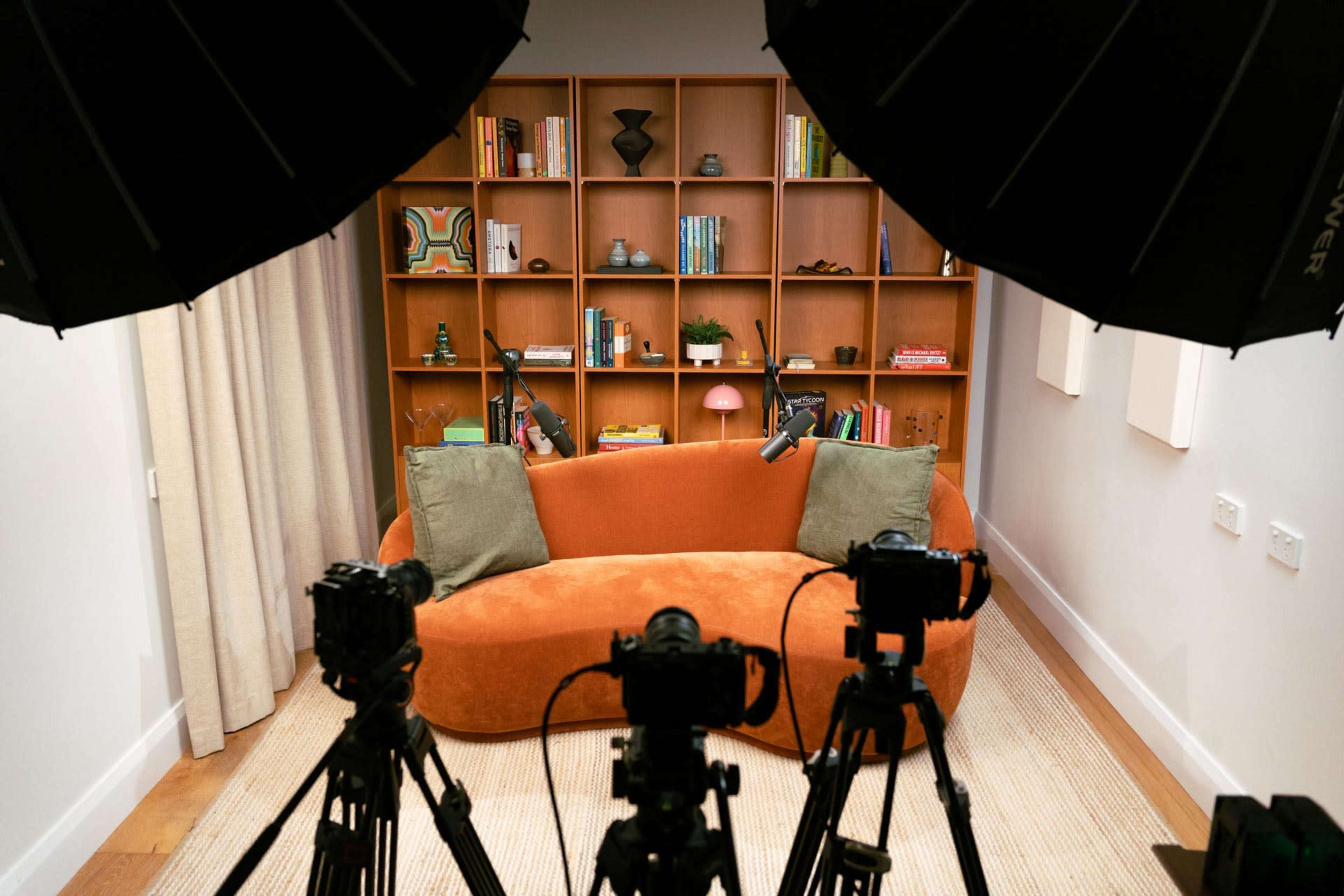 The image shows a cozy orange couch in a well-lit room, with a bookshelf filled with various decor items and books in the background, framed by two large studio lights and several cameras facing the couch.