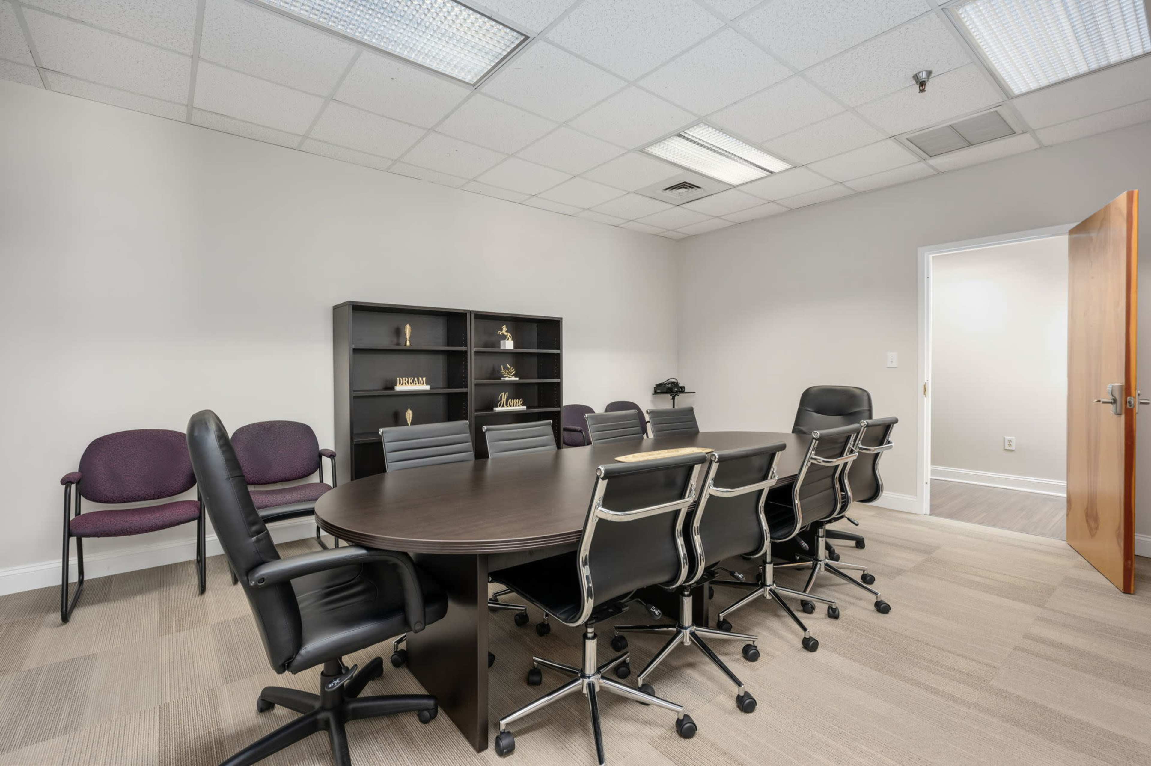 A conference room features a large oval table surrounded by black leather chairs, with a shelving unit against the wall and several empty chairs at one side.