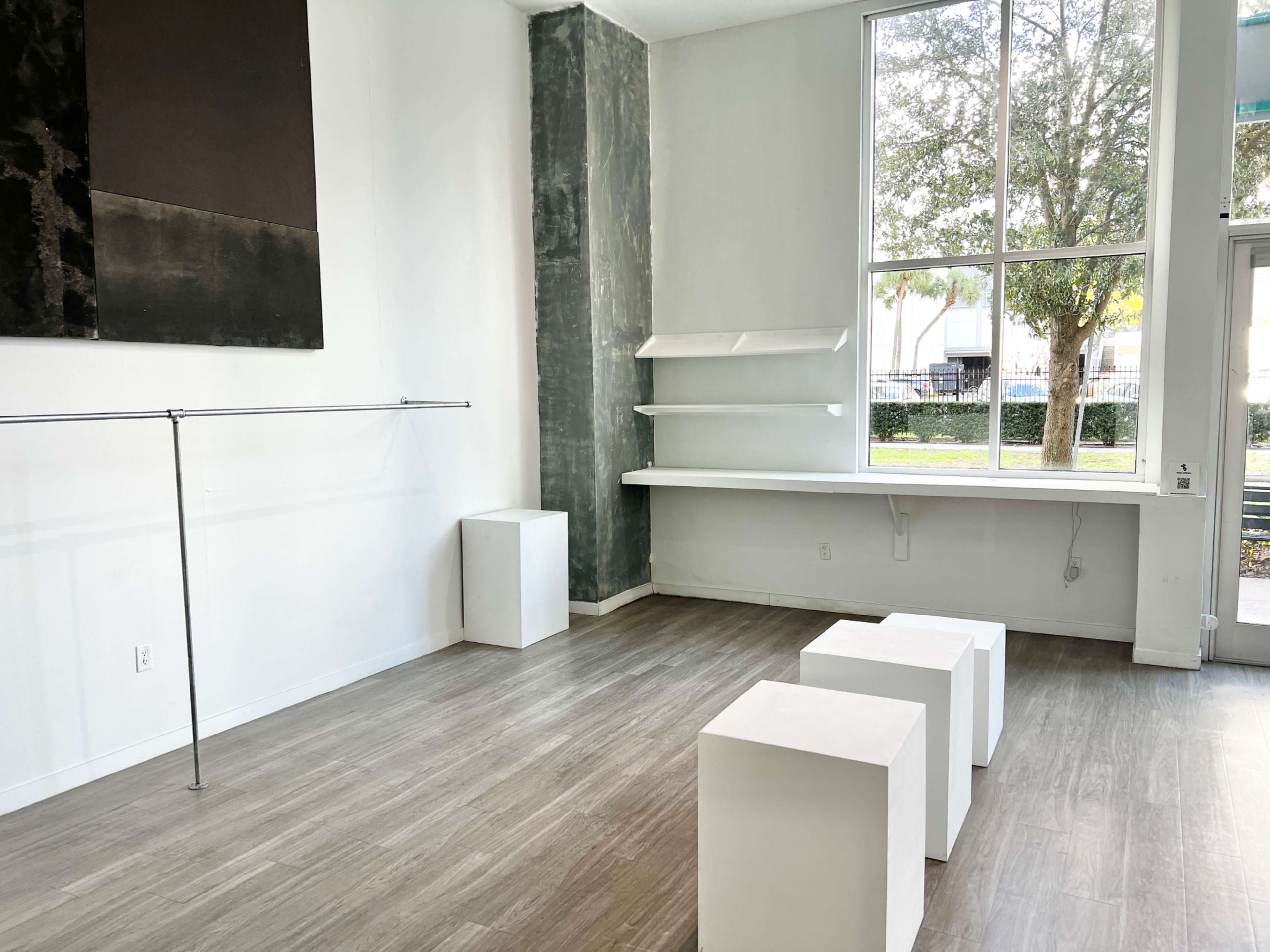 A minimalistic interior with white walls, a gray section, a clothing rack, three white pedestals, and a window revealing greenery outside.