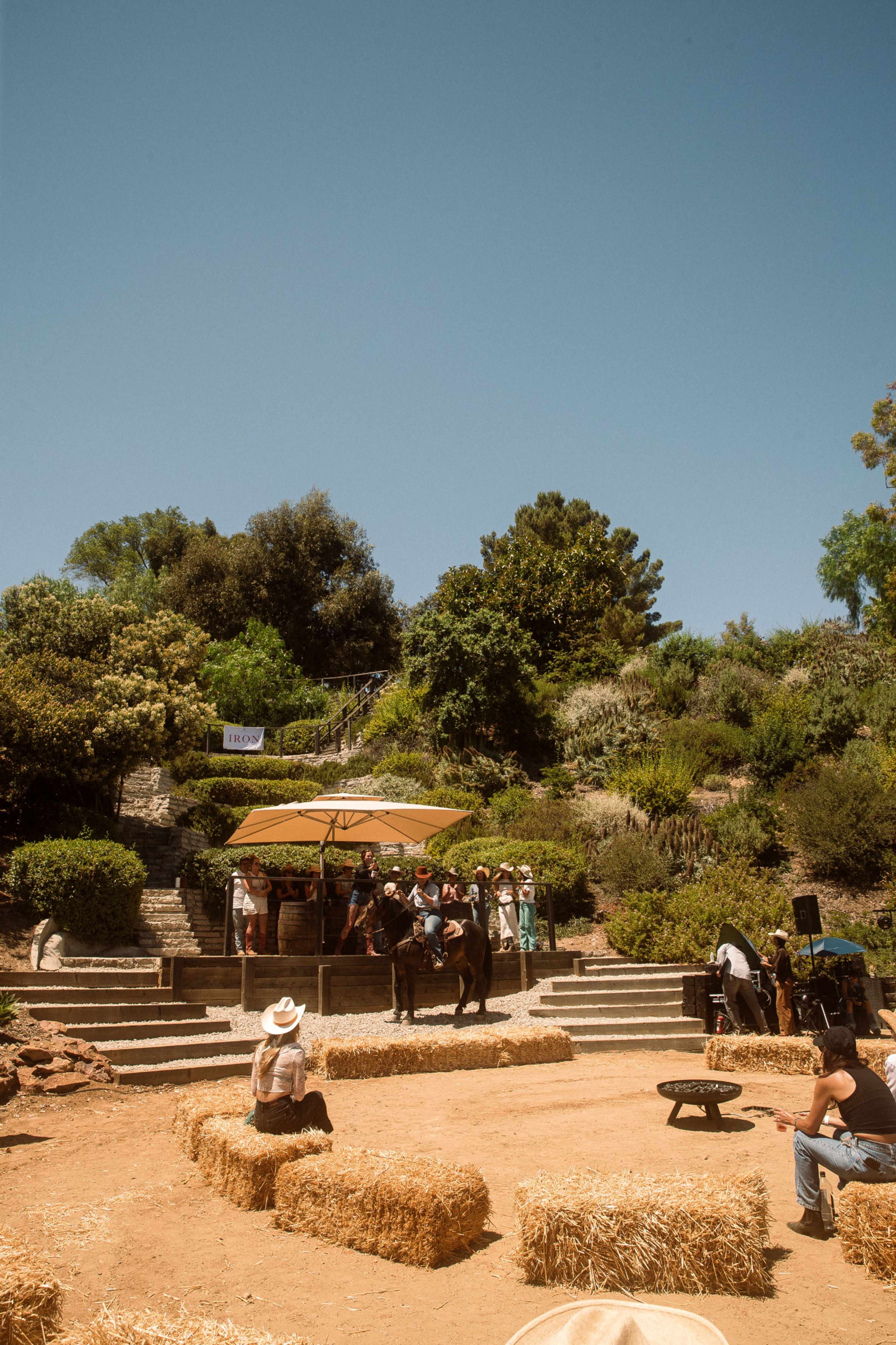 A small audience sits on hay bales in an outdoor setting, watching a performance on a stage beneath an umbrella surrounded by greenery.