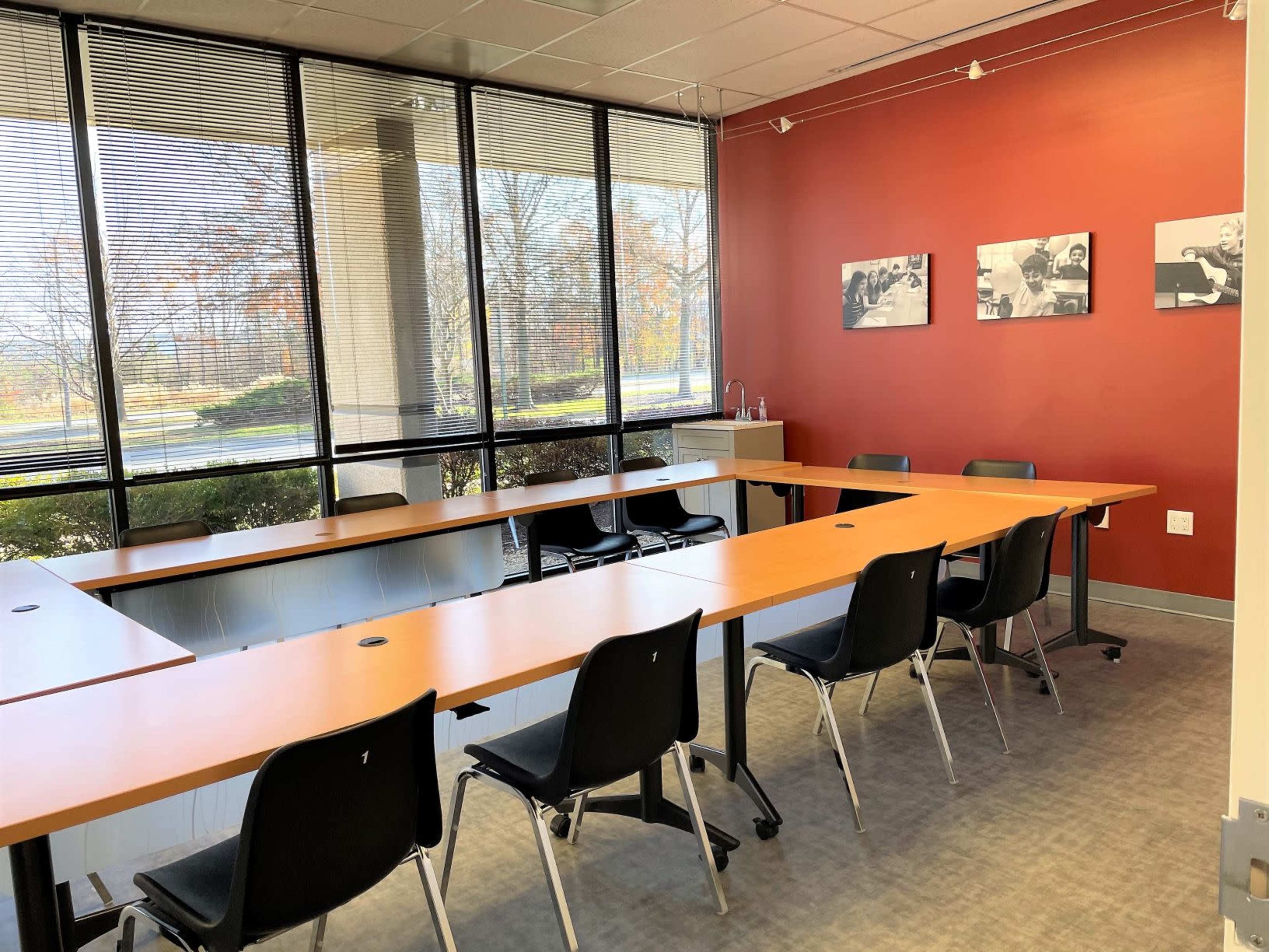 A conference room with a long rectangular table surrounded by black chairs, large windows allowing natural light, and a red accent wall with framed black-and-white photos.