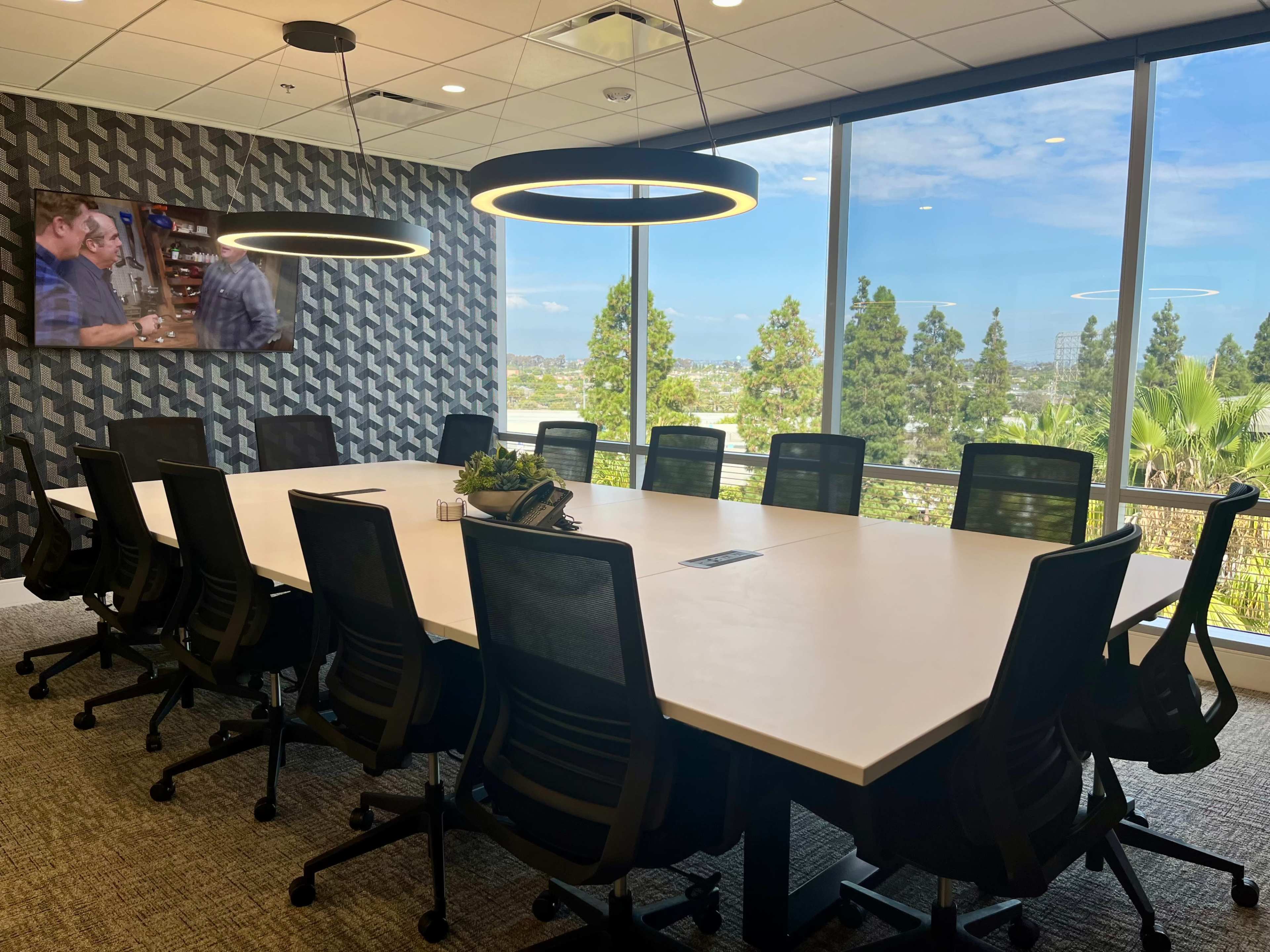 The image shows a modern conference room with a large rectangular table, black ergonomic chairs, a wall-mounted screen, and large windows overlooking trees and a blue sky.