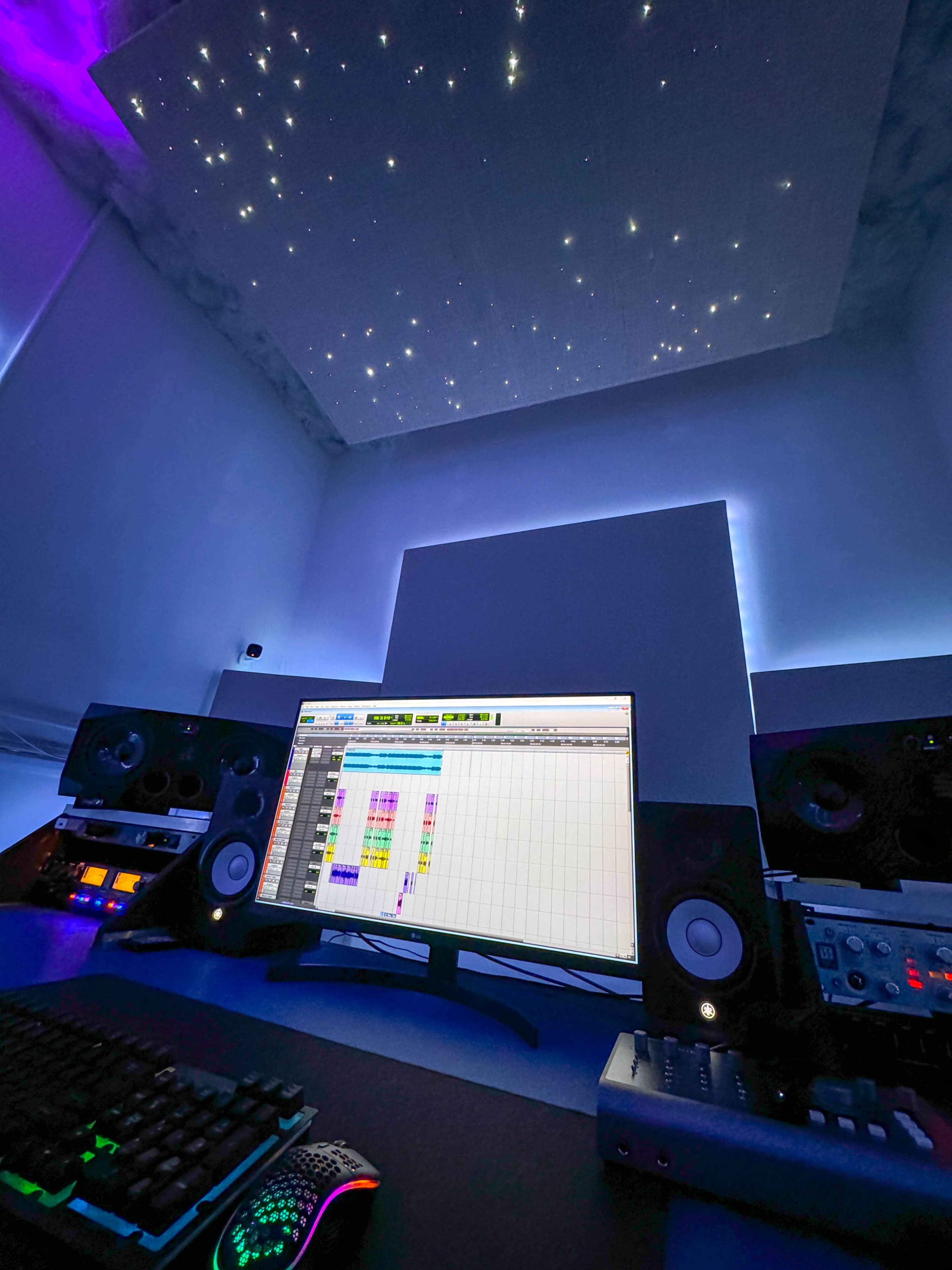 A music production studio features a computer monitor displaying audio tracks, flanked by speakers and a keyboard, with a starry ceiling above.
