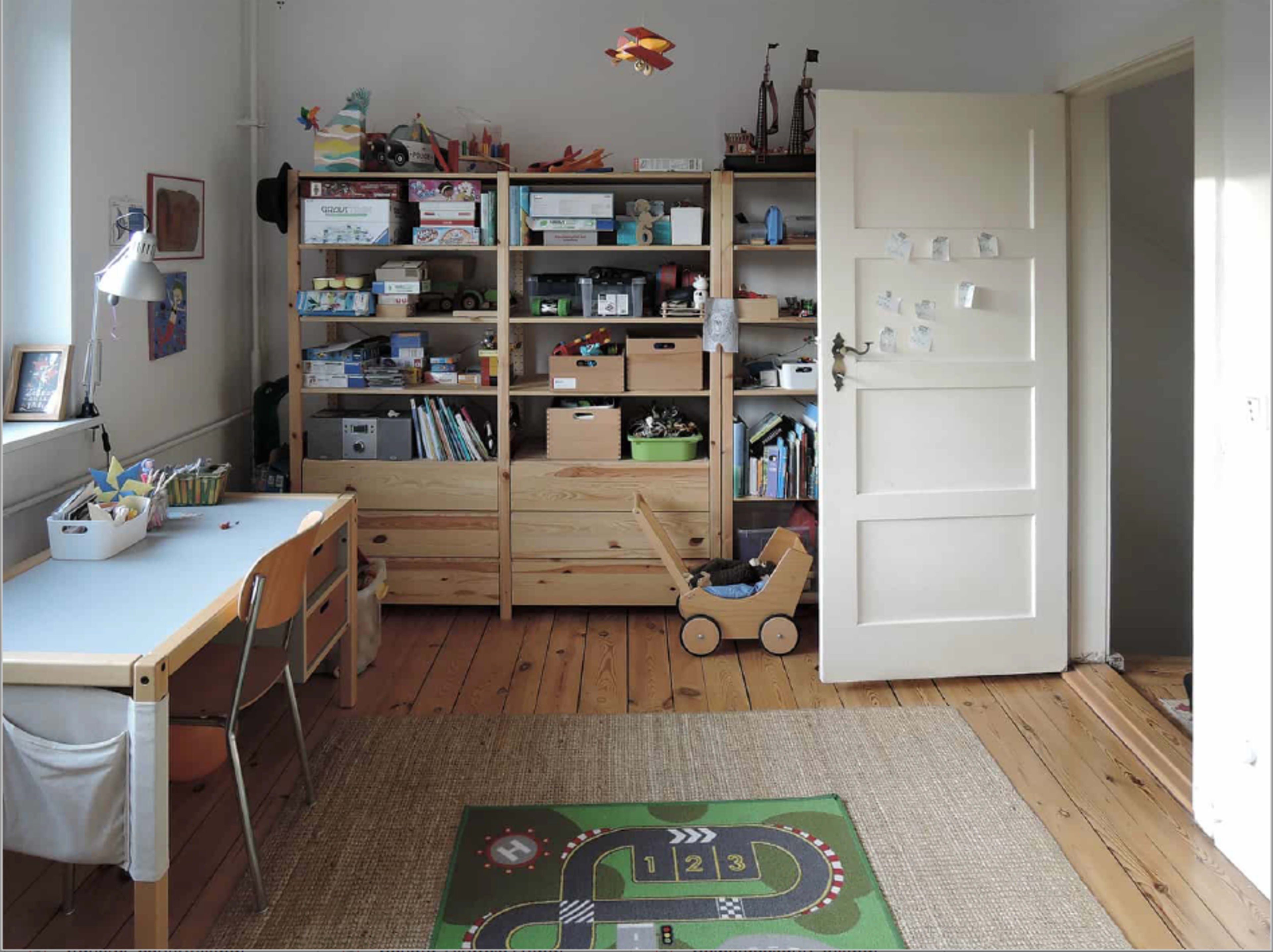 A tidy children's room featuring a wooden shelving unit filled with toys and books, a desk with chairs, and a toy cart on a patterned rug.