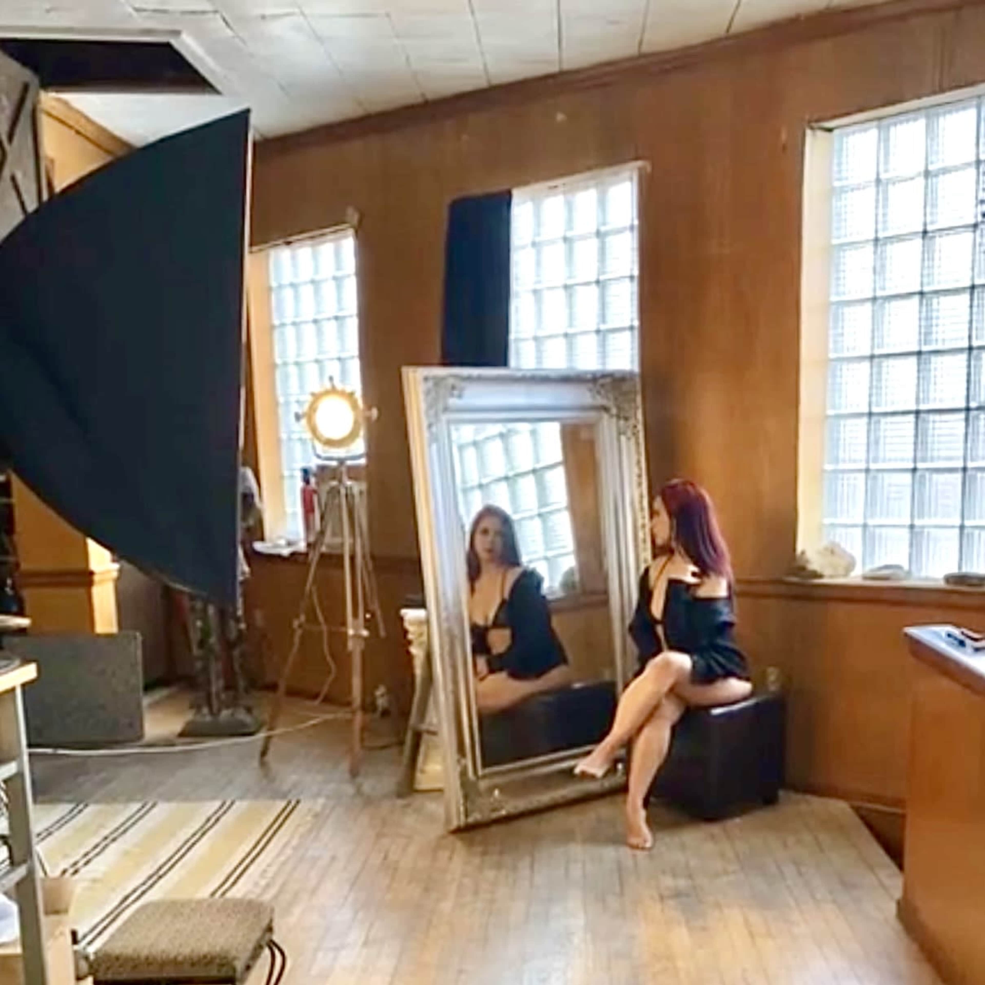 A model poses in front of a large mirror in a well-lit studio with wooden walls and large windows.