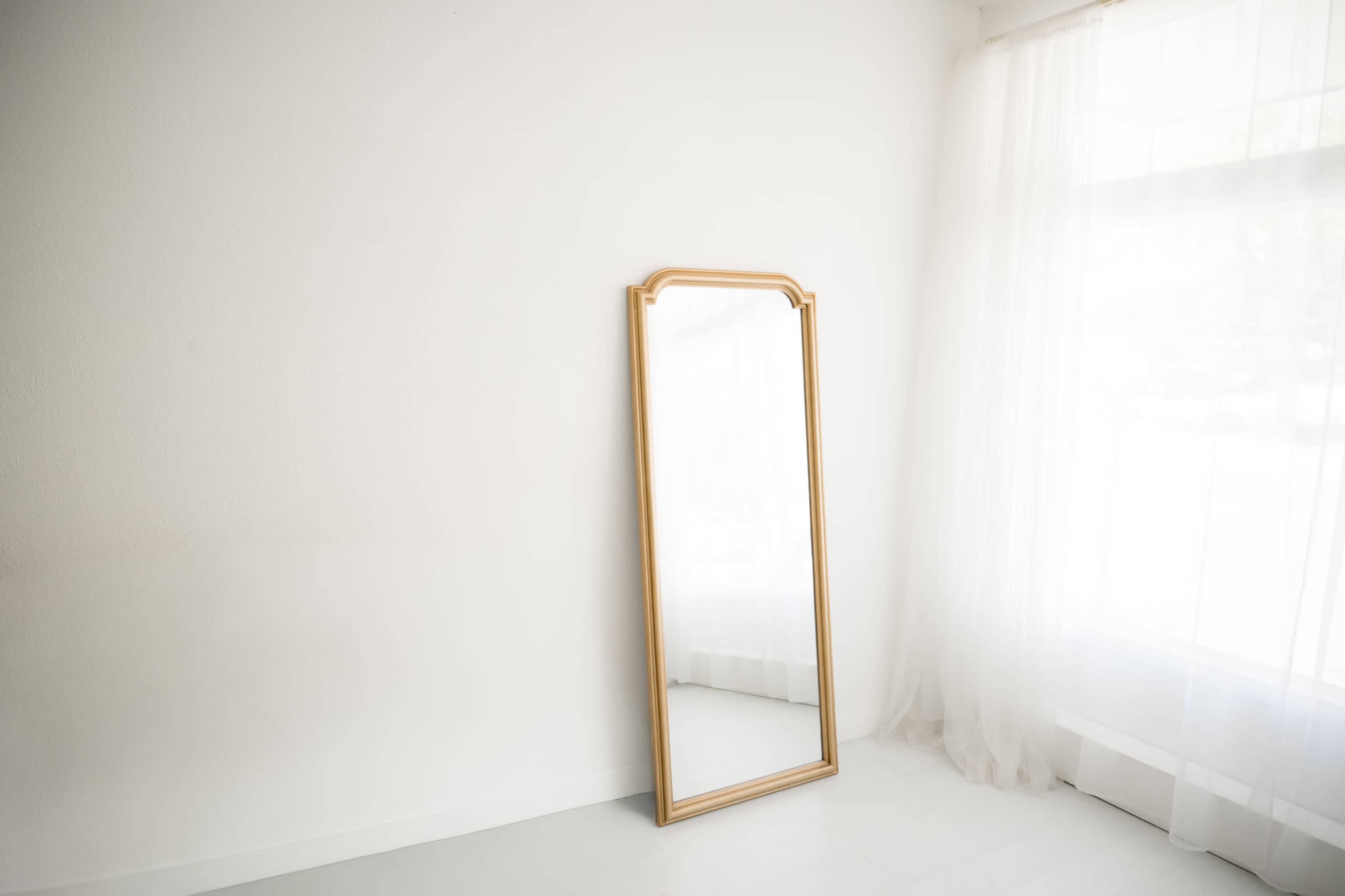 A tall, wooden-framed mirror stands against a white wall near a window draped with sheer curtains.
