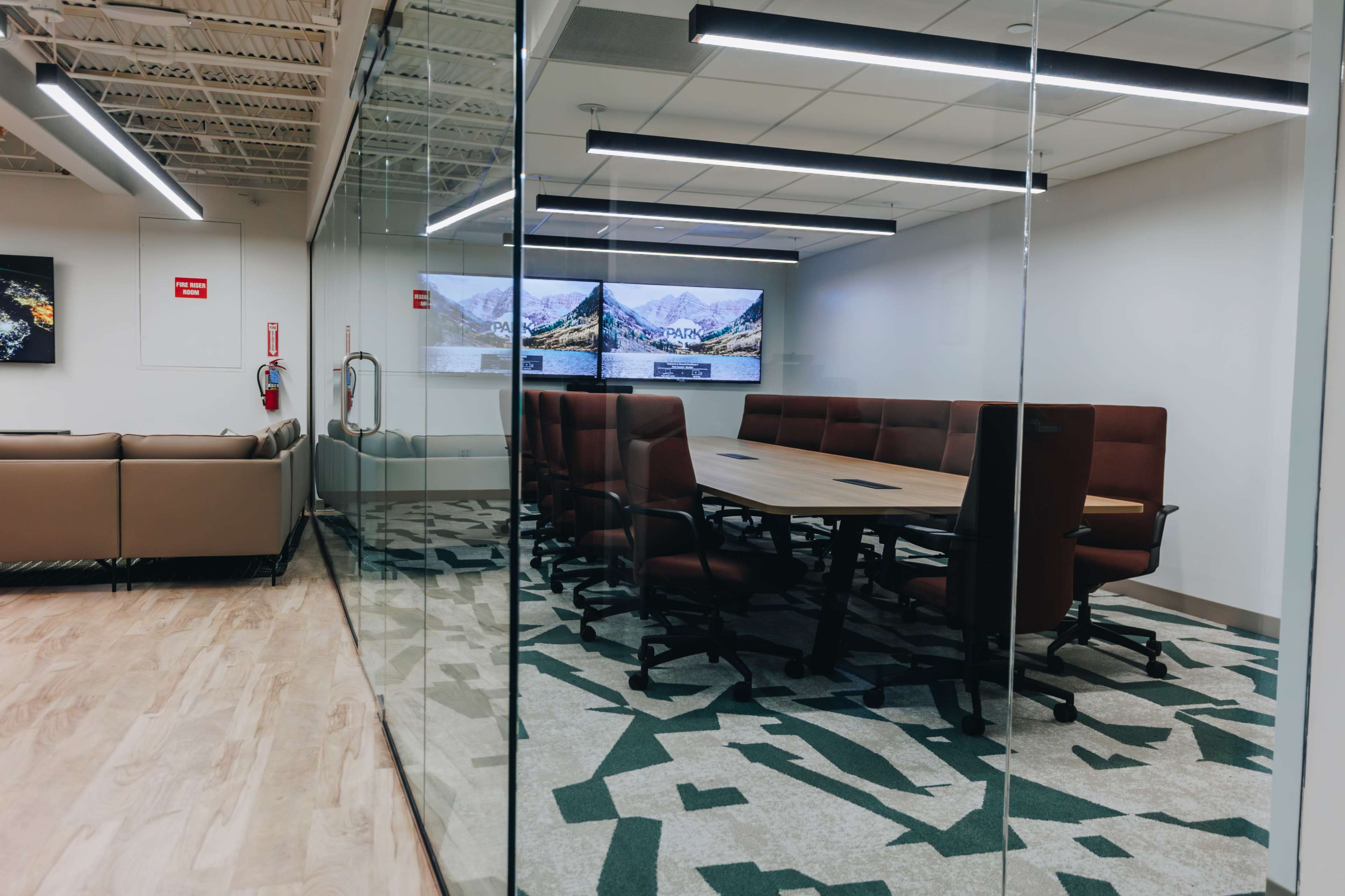 The image shows a modern conference room with a large rectangular table surrounded by brown swivel chairs, glass walls, and a scenic digital display on the back wall.