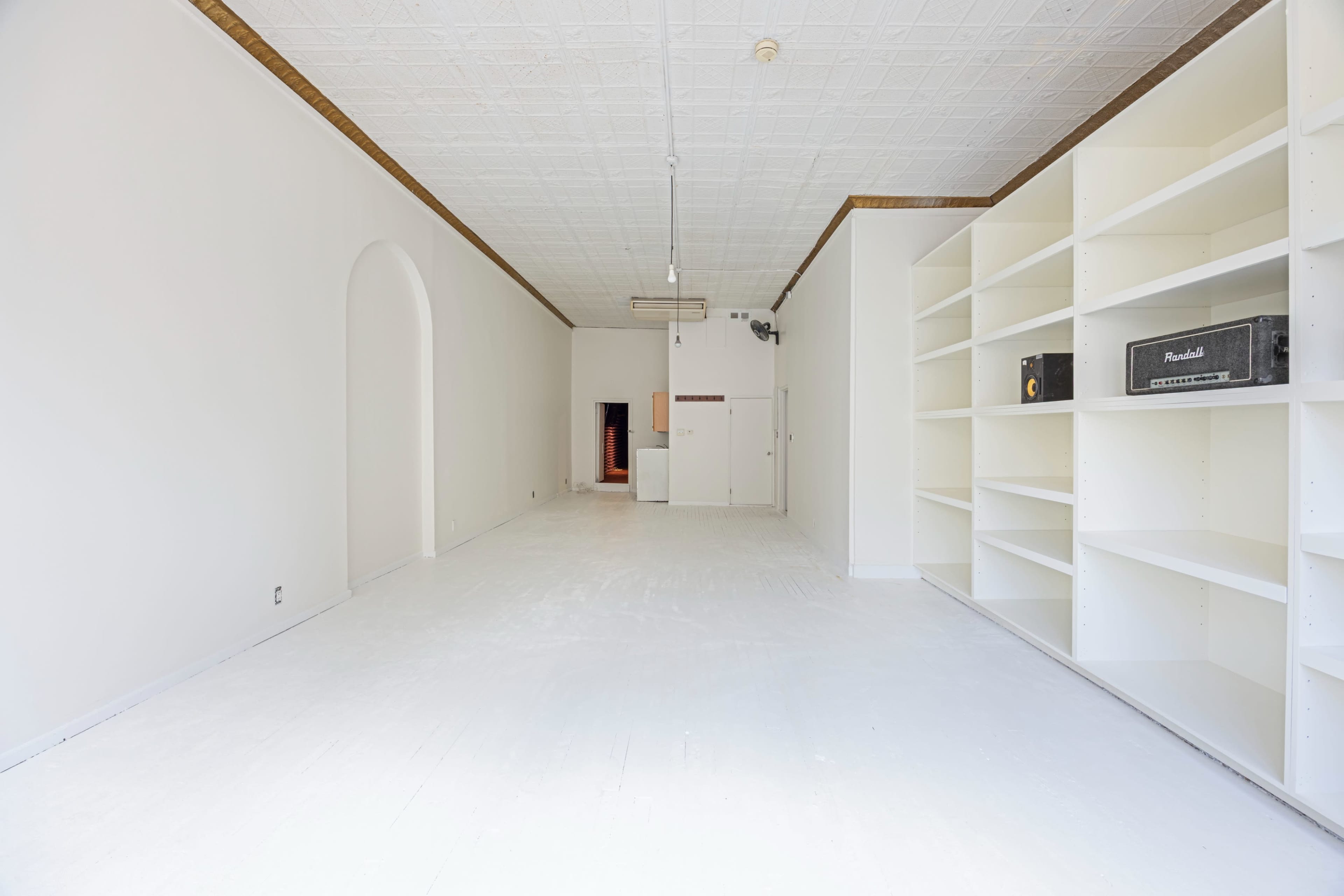 The image shows an empty, well-lit room with white walls and flooring, featuring a shelf unit on the right and an archway on the left.