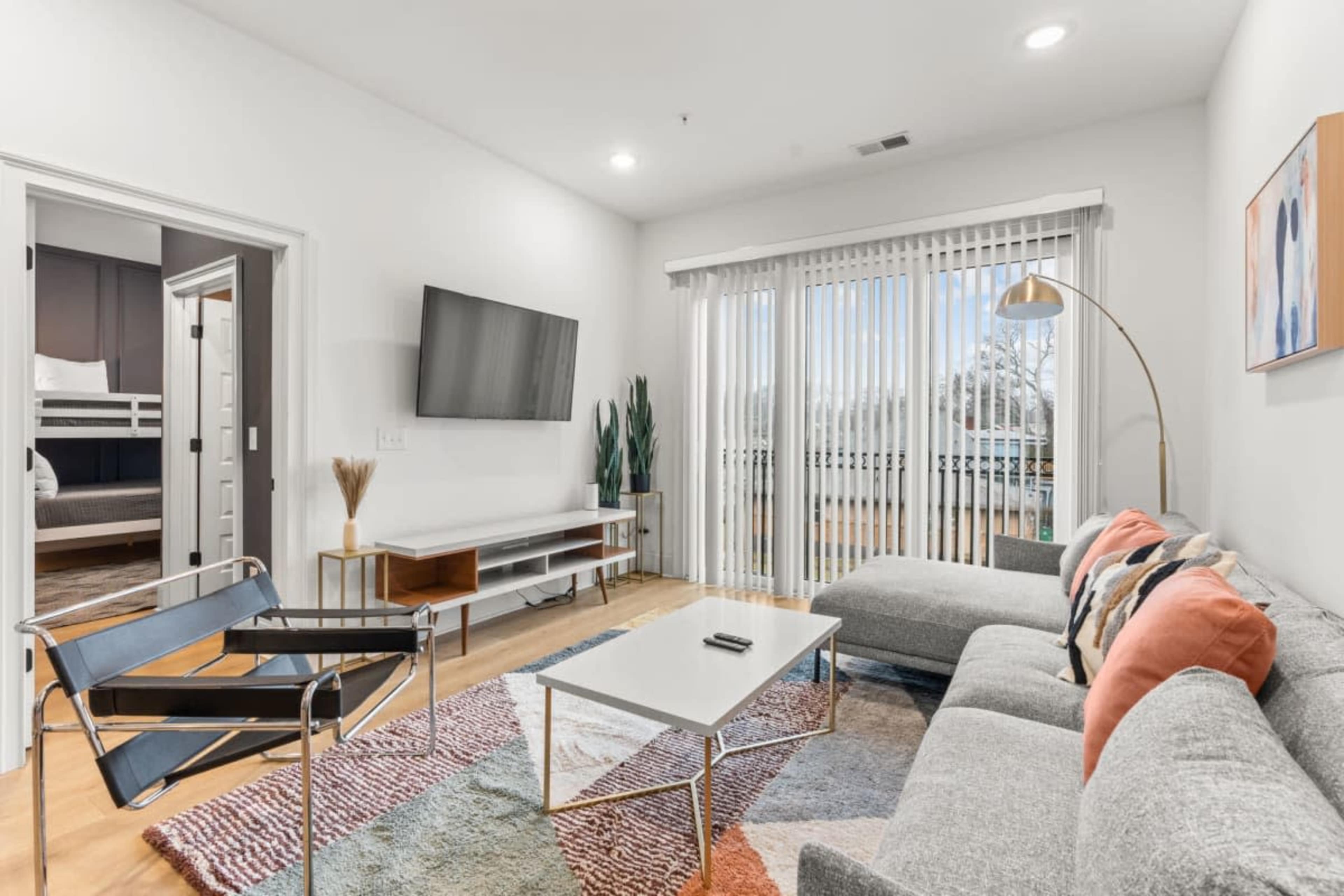 A modern living room with a sectional sofa, a coffee table, a flat-screen TV on the wall, and a sliding glass door leading to a balcony.