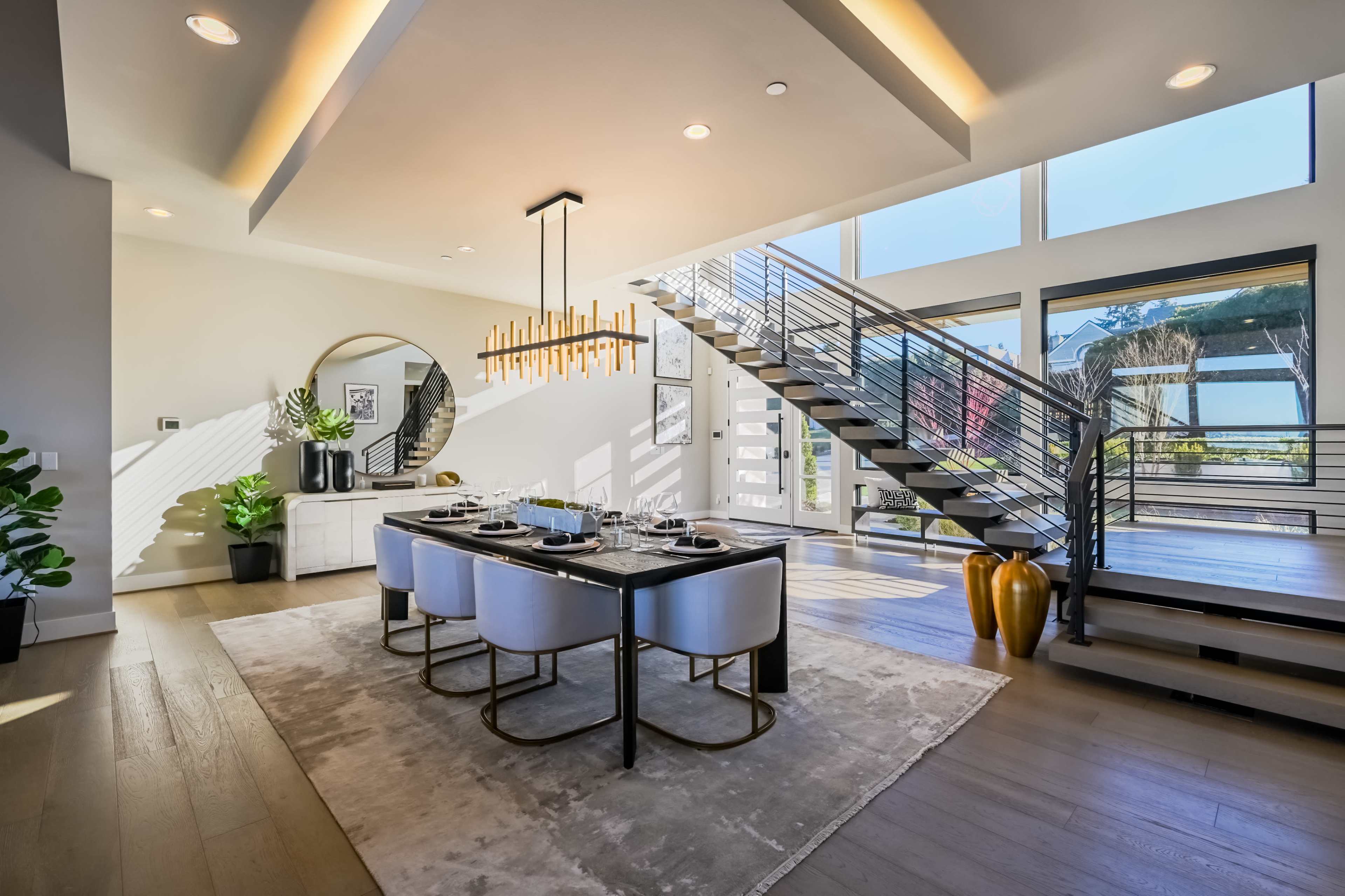 A modern dining area with a large table surrounded by white chairs, a spiral staircase, and large windows allowing natural light in.