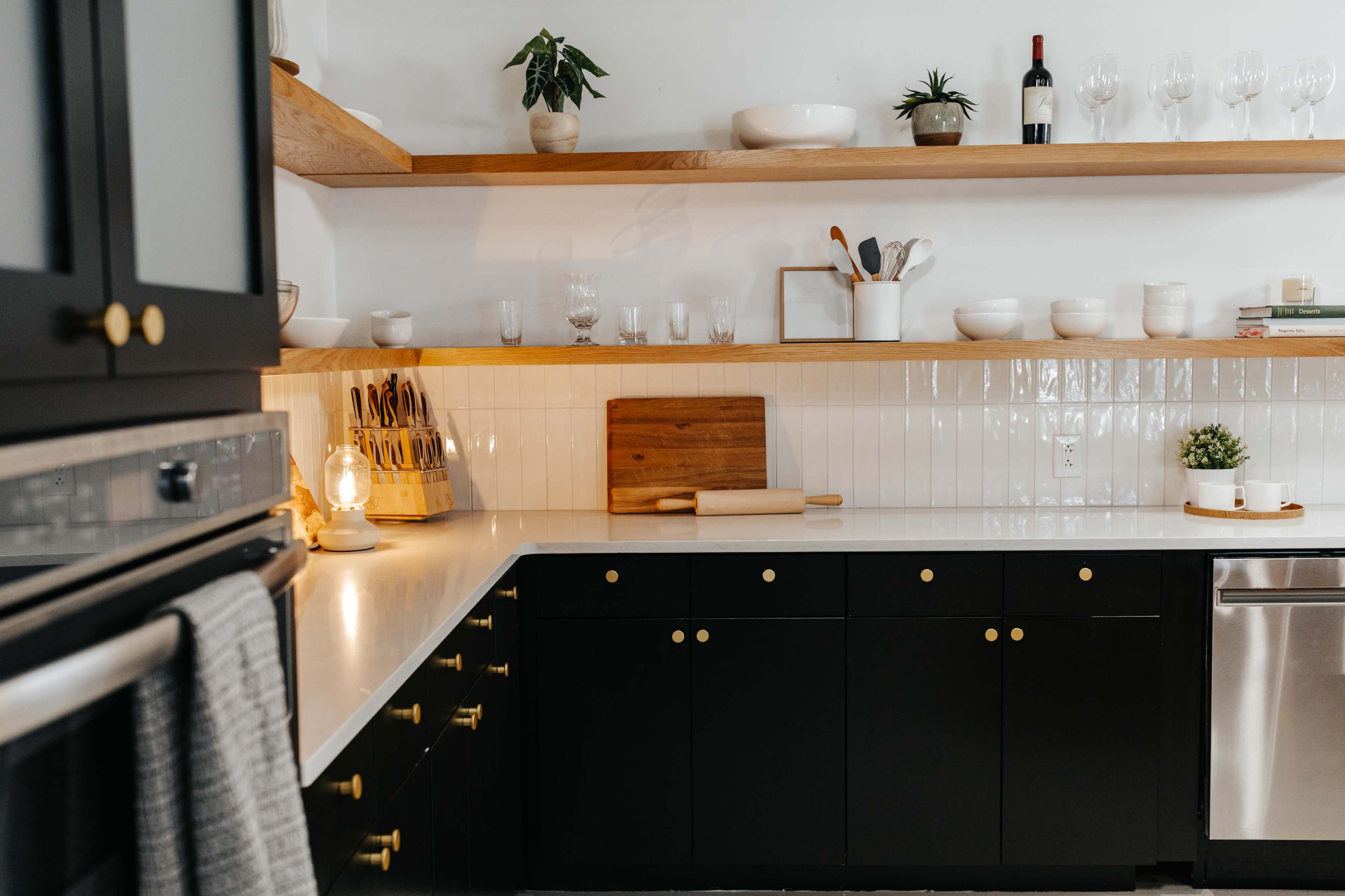 A modern kitchen features black cabinetry, white counters, open shelving with plants, glassware, and a wood cutting board.
