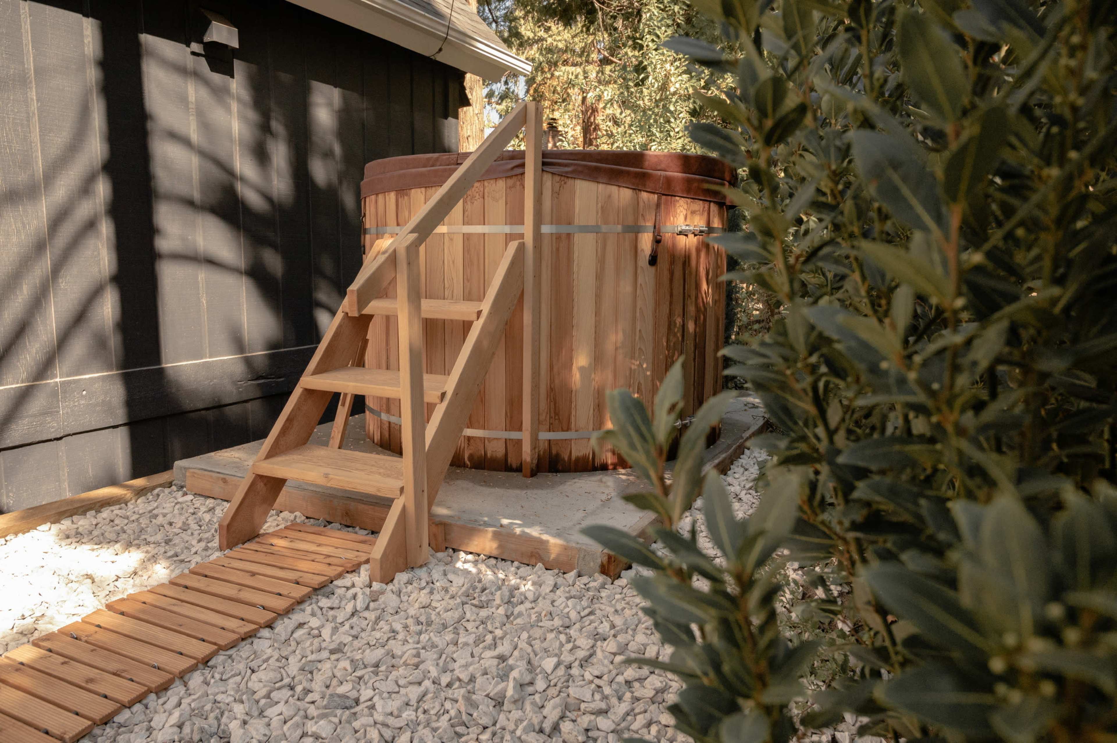 A wooden hot tub with steps is positioned among gravel and greenery outside a building.