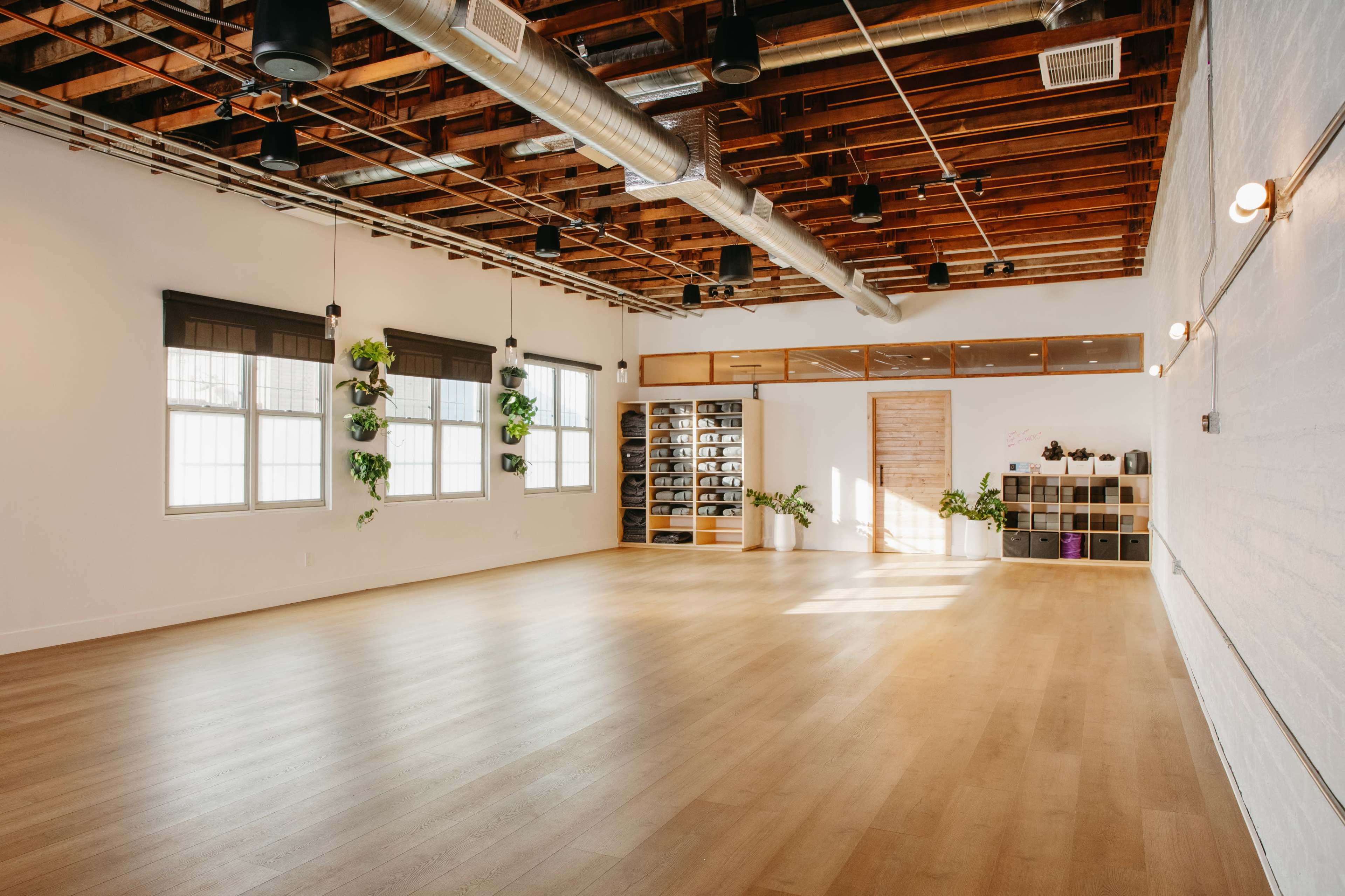 The image shows a spacious, well-lit studio with wooden floors, exposed beams, and large windows adorned with plants.