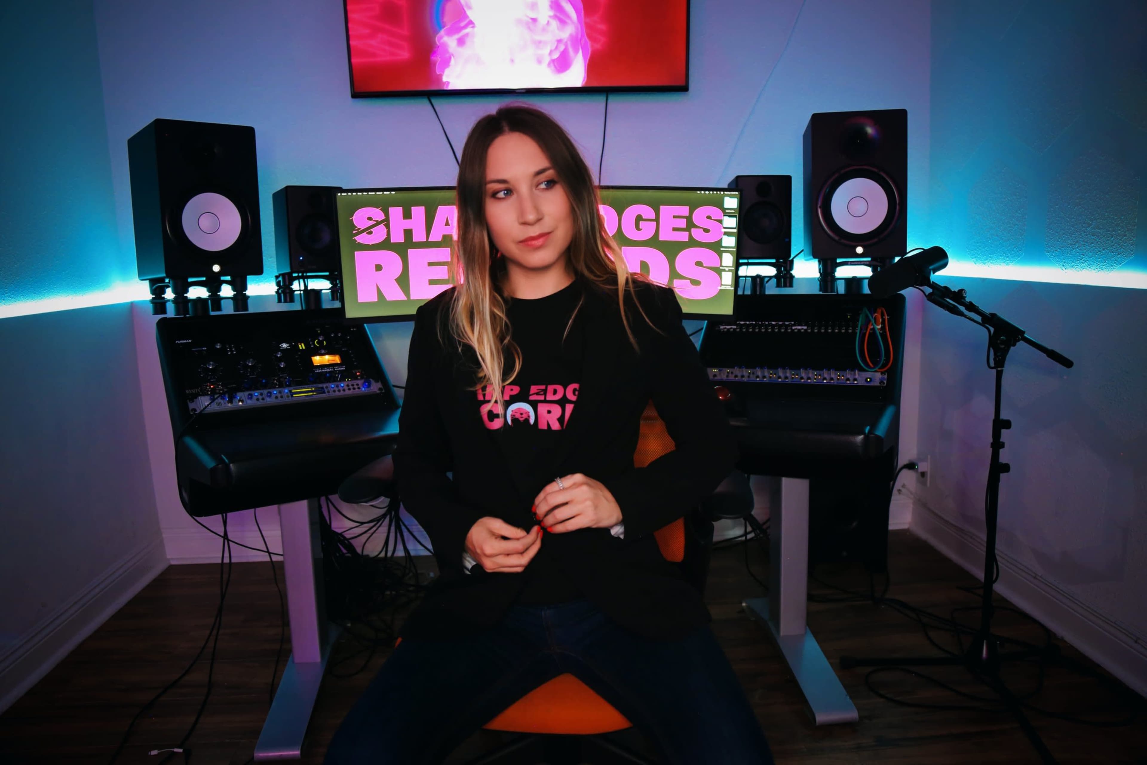 A woman sits in a chair in a music studio surrounded by audio equipment and monitors, with a colorful screen displaying "SHARP EDGES RECORDS" in the background.