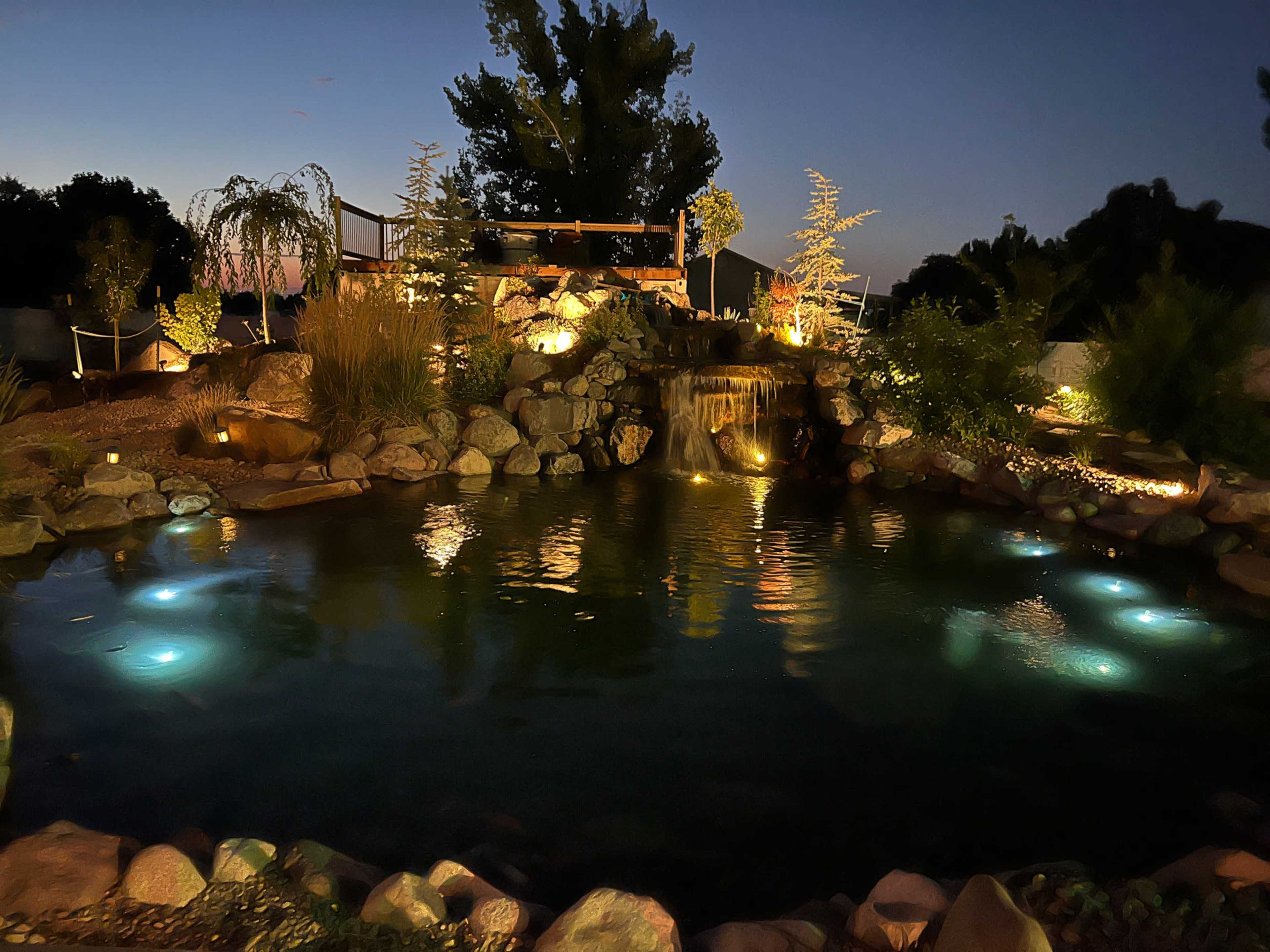 A serene pond with illuminated water features and surrounding rocks is set against a twilight sky, featuring lush greenery and a small waterfall.