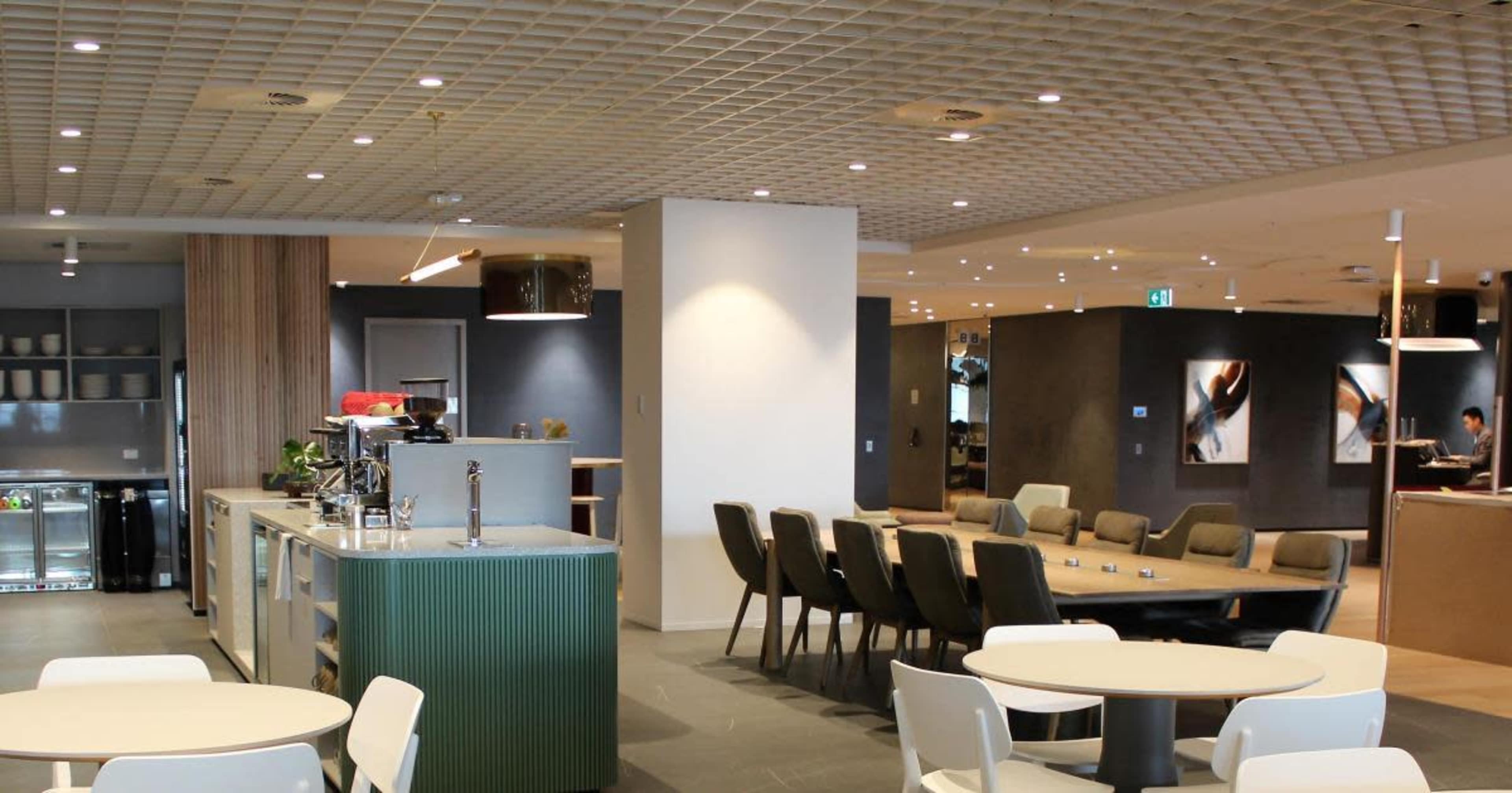 The image shows a modern lounge area with tables and chairs, a counter with a beverage dispenser, and artwork on the walls.