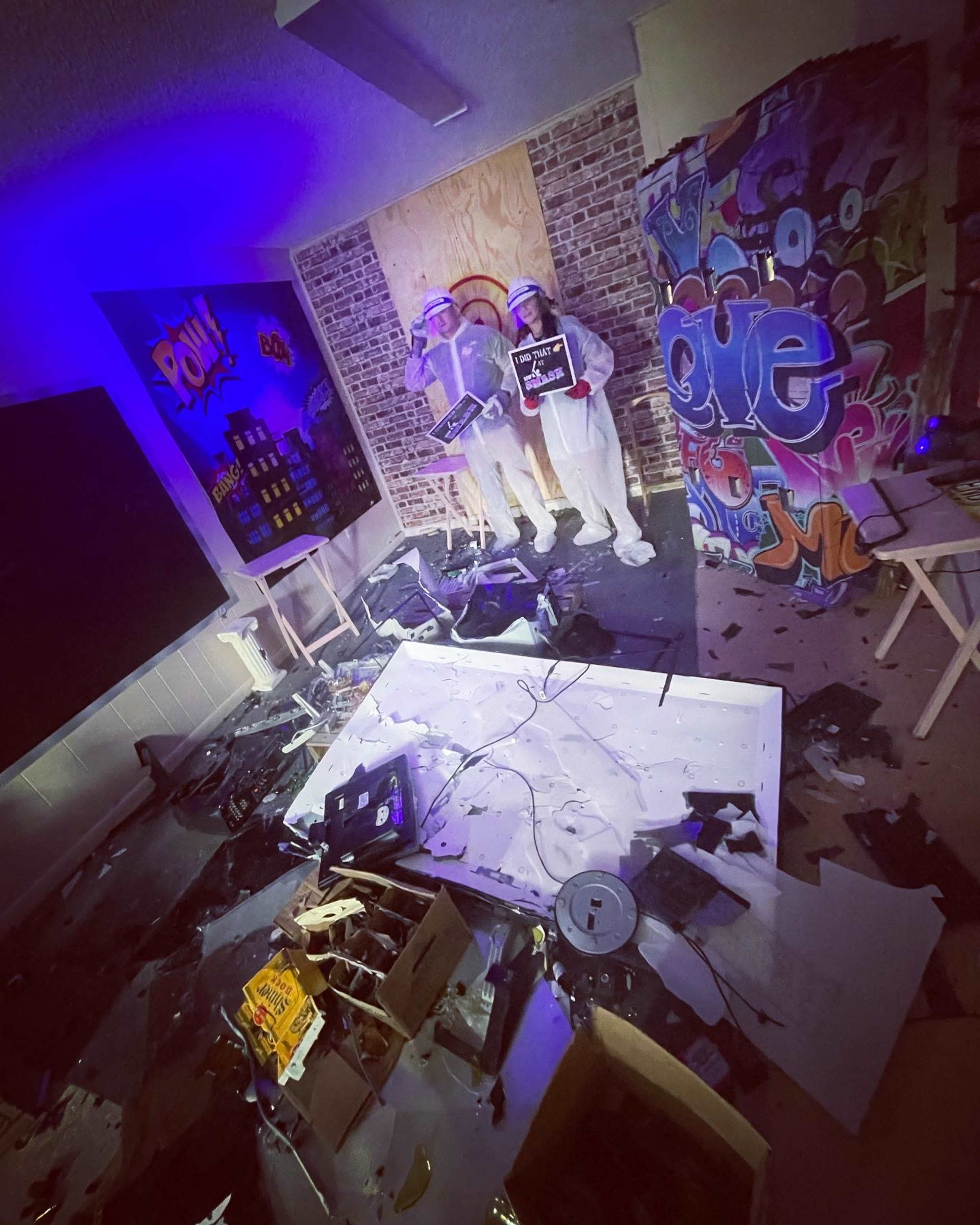 The image depicts a cluttered room with scattered debris, including broken electronics and art supplies, alongside two figures dressed in white protective clothing holding signs.