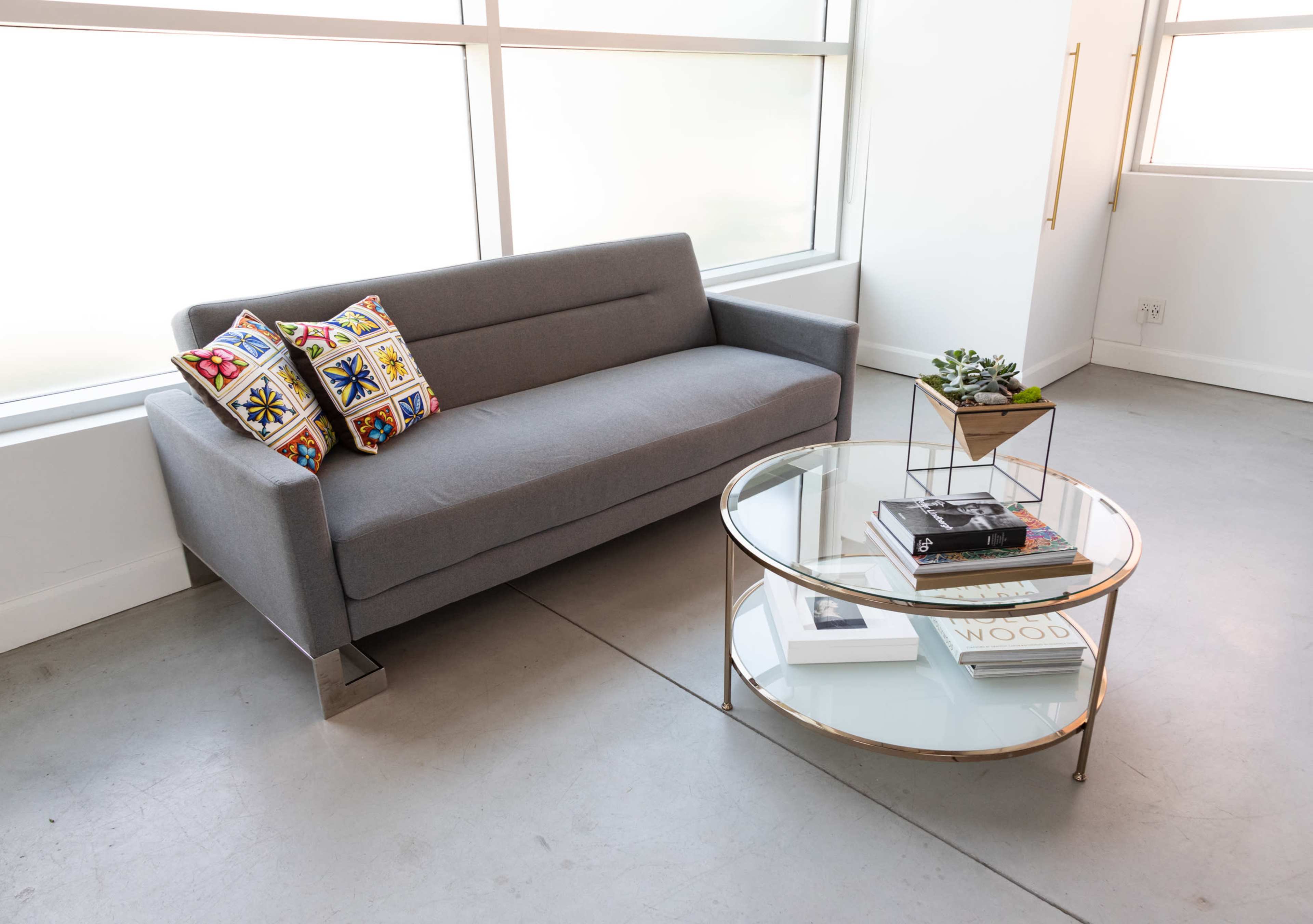 A gray sofa with decorative pillows is positioned next to a round glass coffee table holding books and a small plant in a modern interior space.