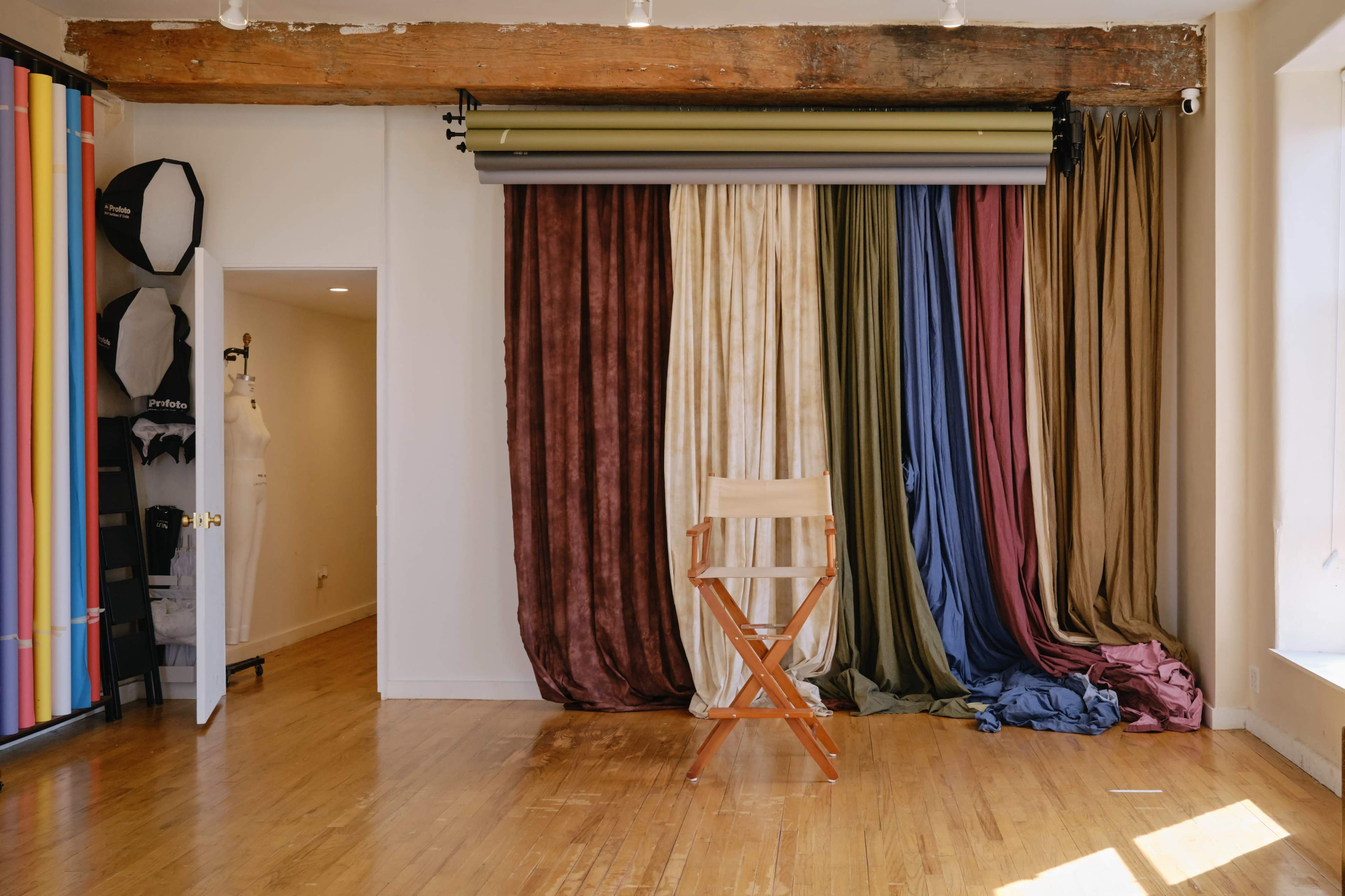 The image shows a photography studio with a wooden floor, featuring a wooden director's chair in front of a backdrop of hanging fabric in various colors.