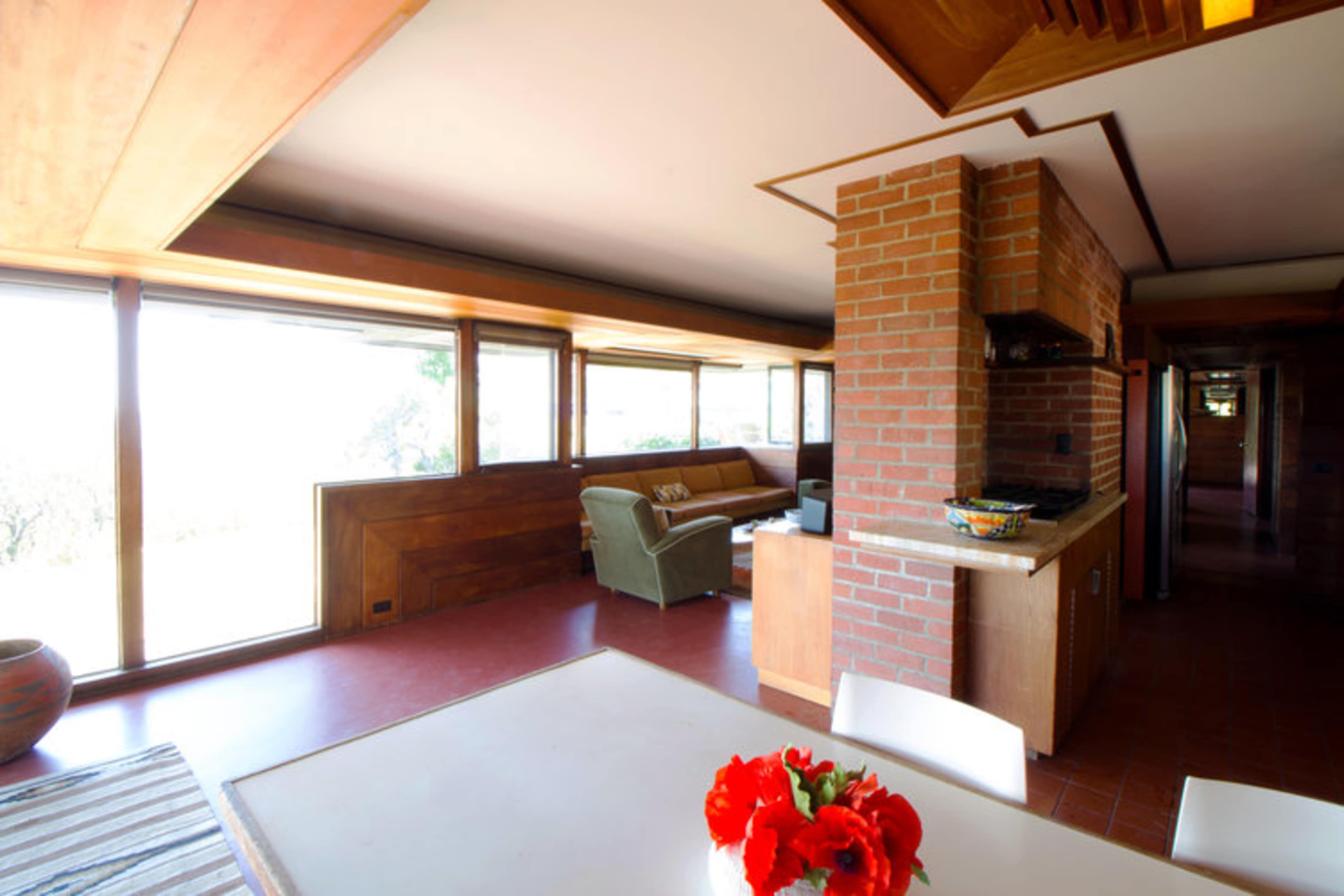 The image shows a spacious interior of a modern house featuring a dining area with a table, a kitchen with brick accents, and large windows providing natural light.