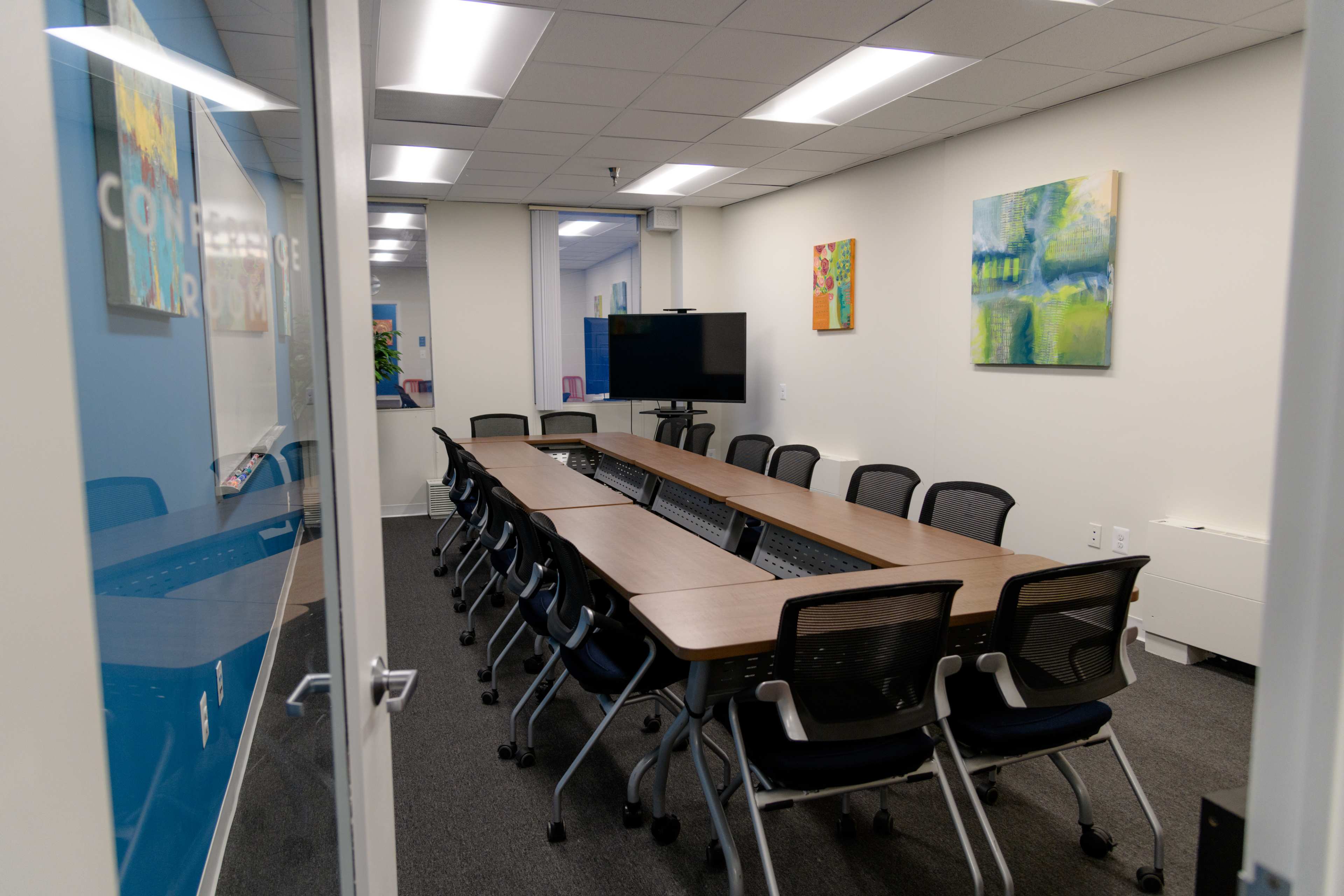 A conference room features a long rectangular table surrounded by chairs, with a television mounted on the wall and colorful artwork displayed.