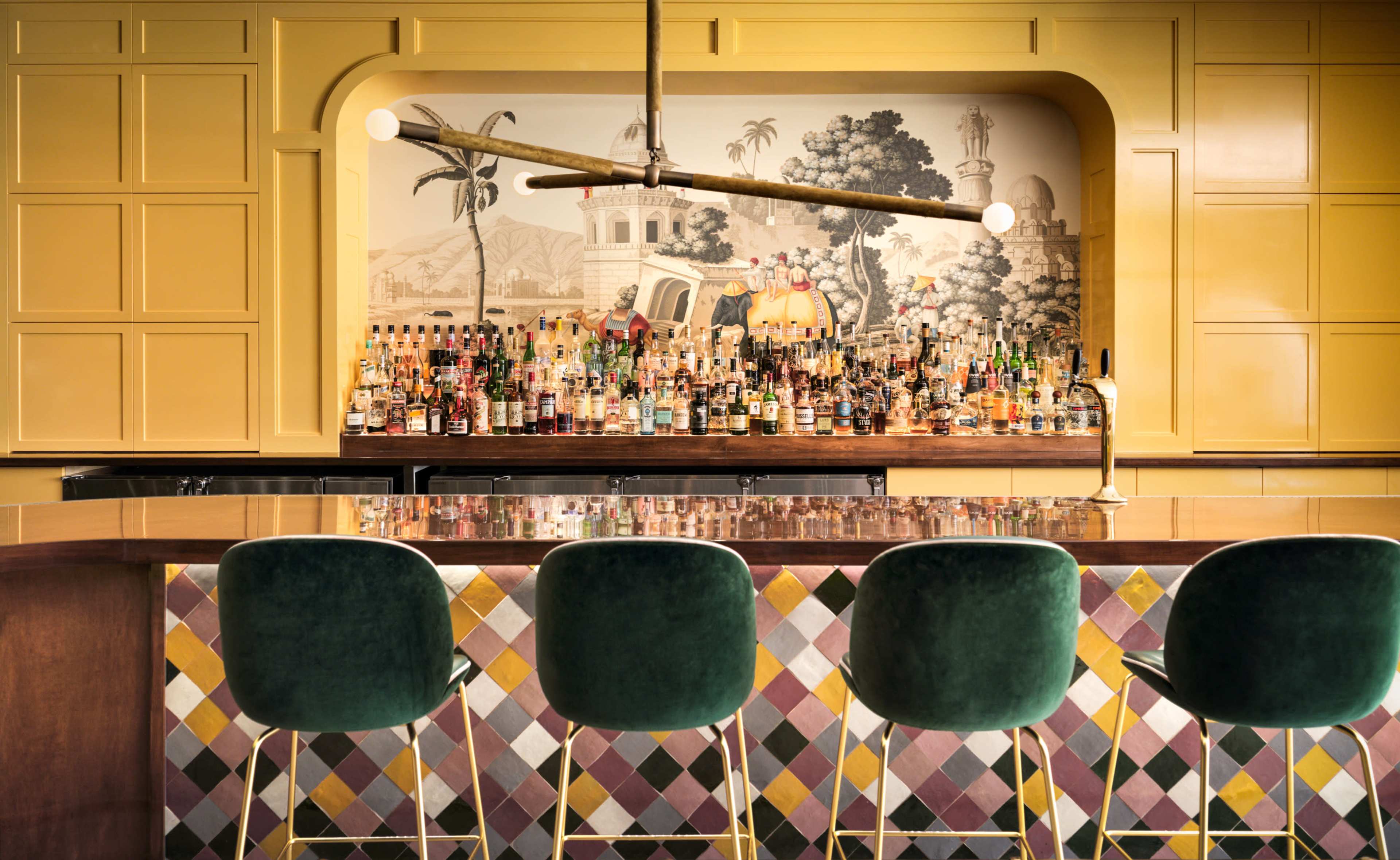 The image shows a stylish bar area with a colorful tiled floor, a wooden counter, and a wall of bottles behind the bar, set against a mural of palm trees and a building.
