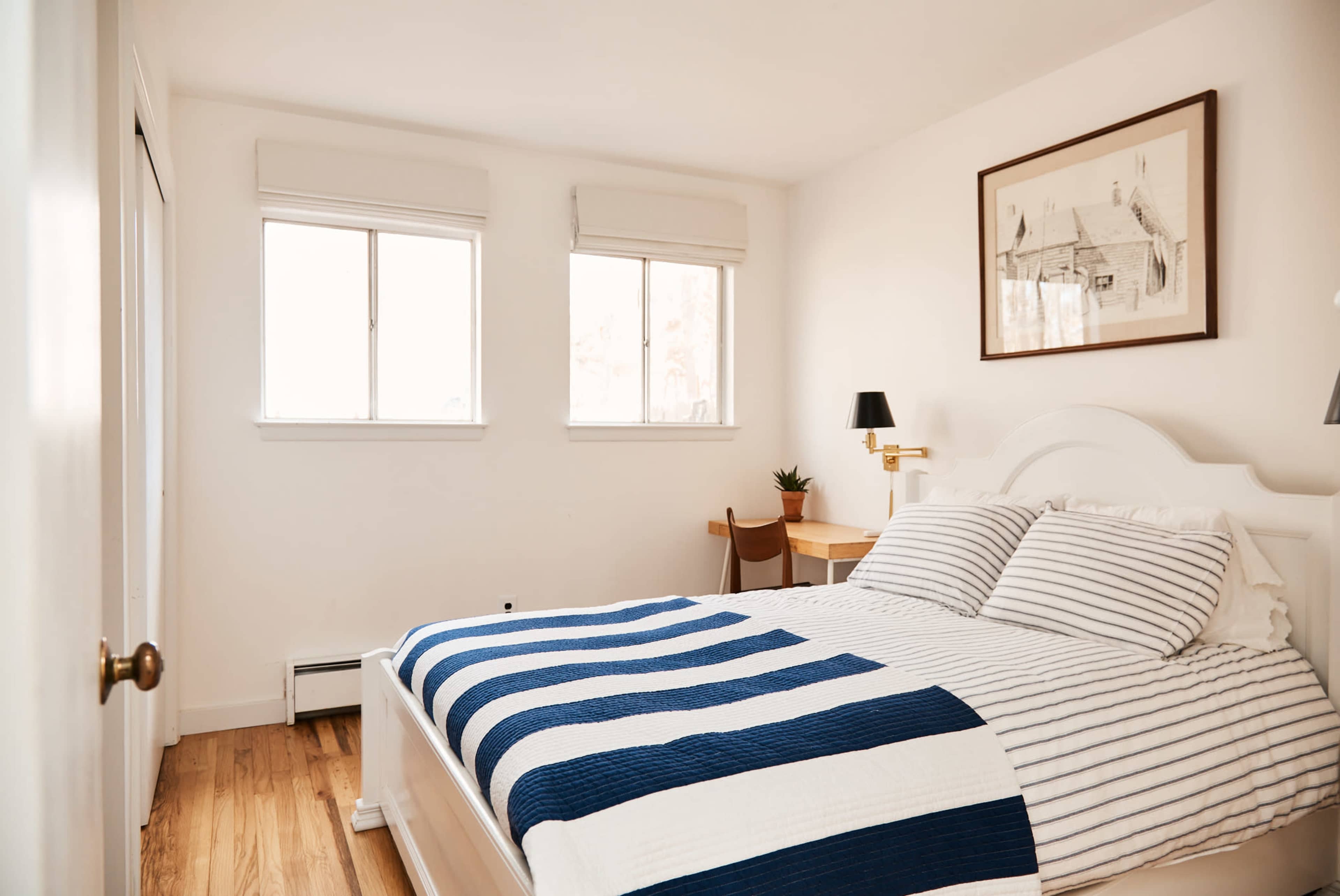 A bright, minimalist bedroom with a white bed dressed in striped bedding, a wooden floor, and two windows allowing natural light to enter.
