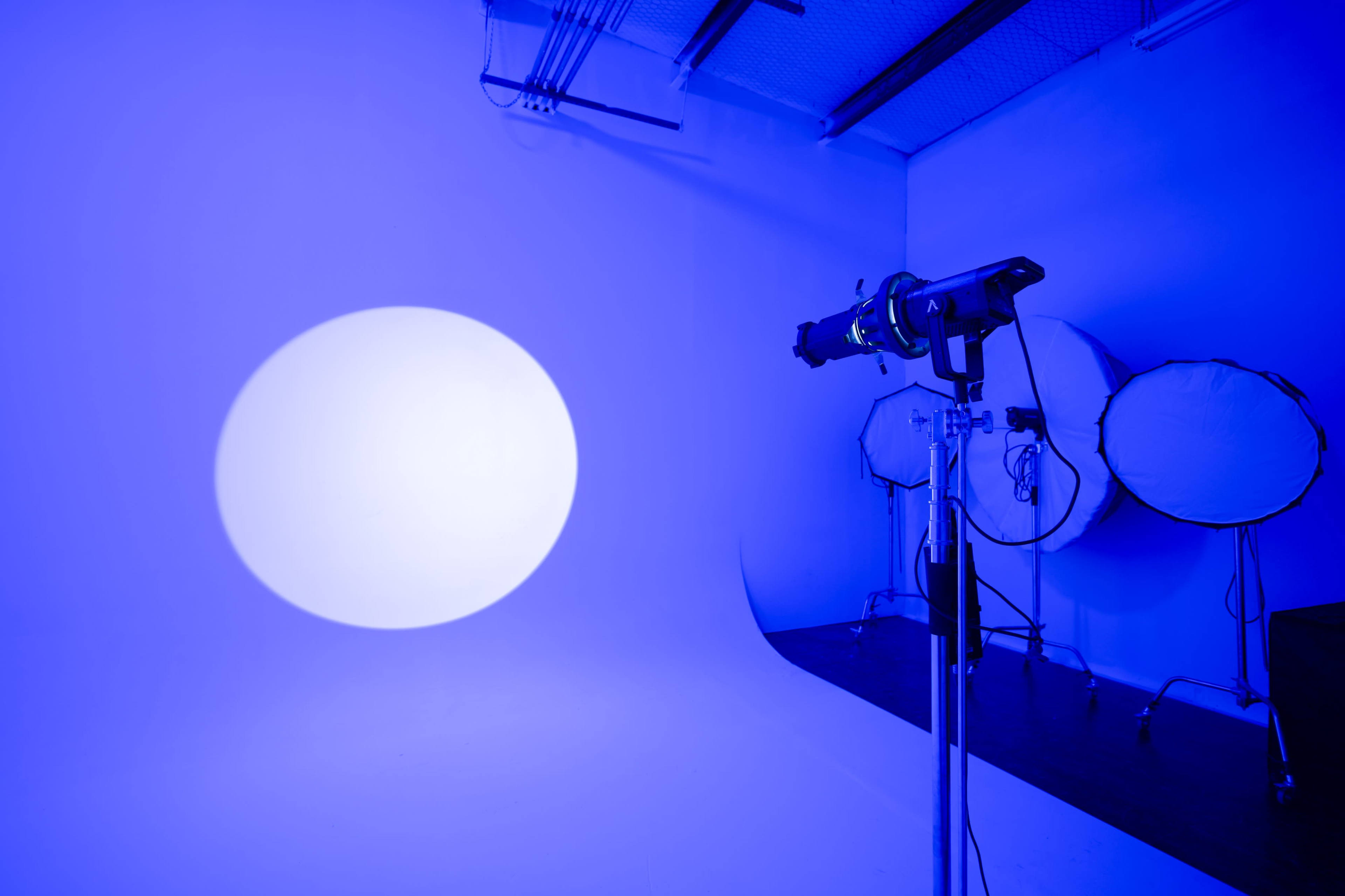 The image shows a photography studio with a large blue light illuminating a circular area on a white backdrop, accompanied by softboxes in the background.