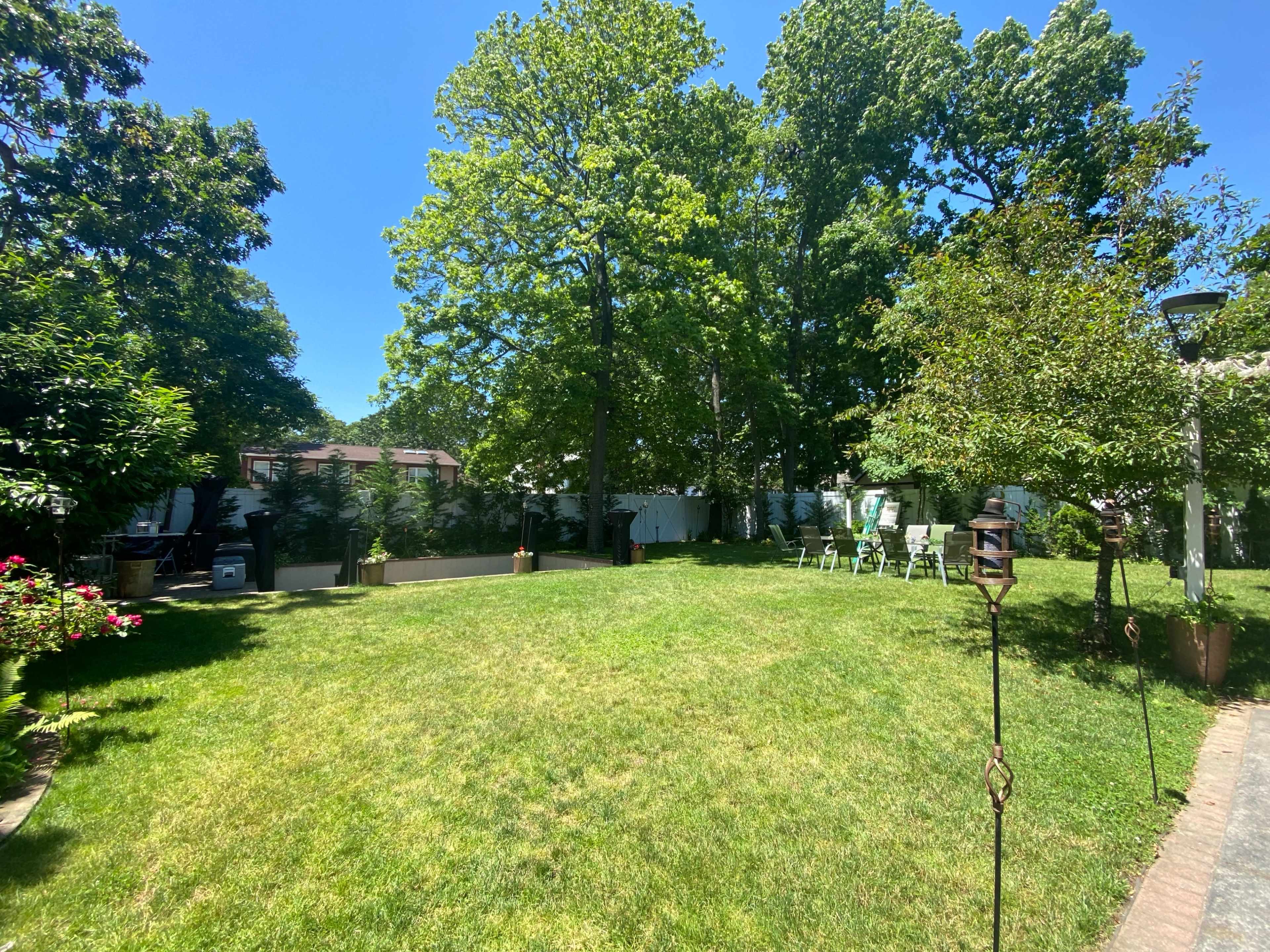 A spacious backyard features green grass, tall trees, and white chairs arranged in the distance under clear blue skies.