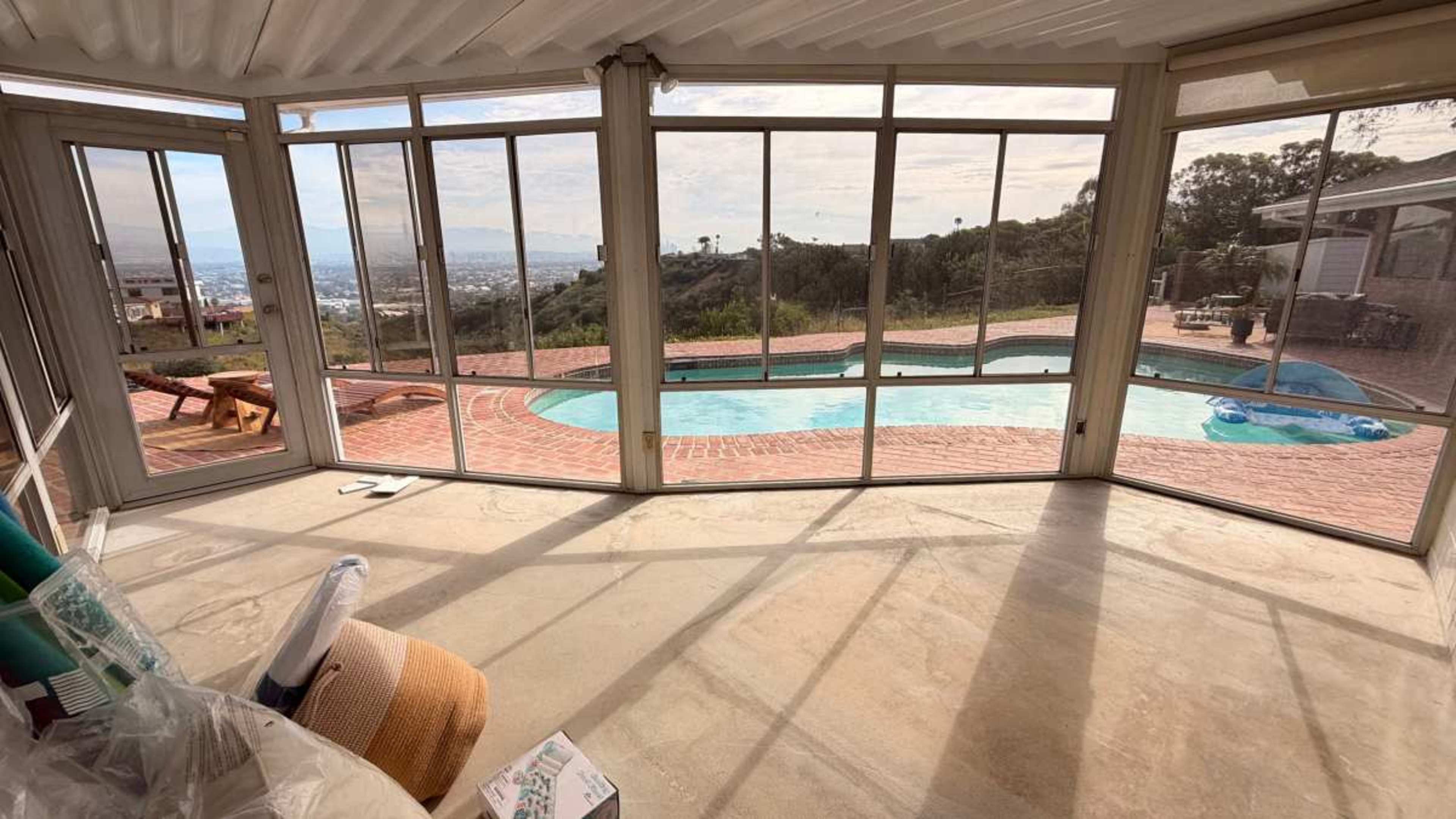 The image shows a spacious sunroom with large windows overlooking a pool and a panoramic view of the city.