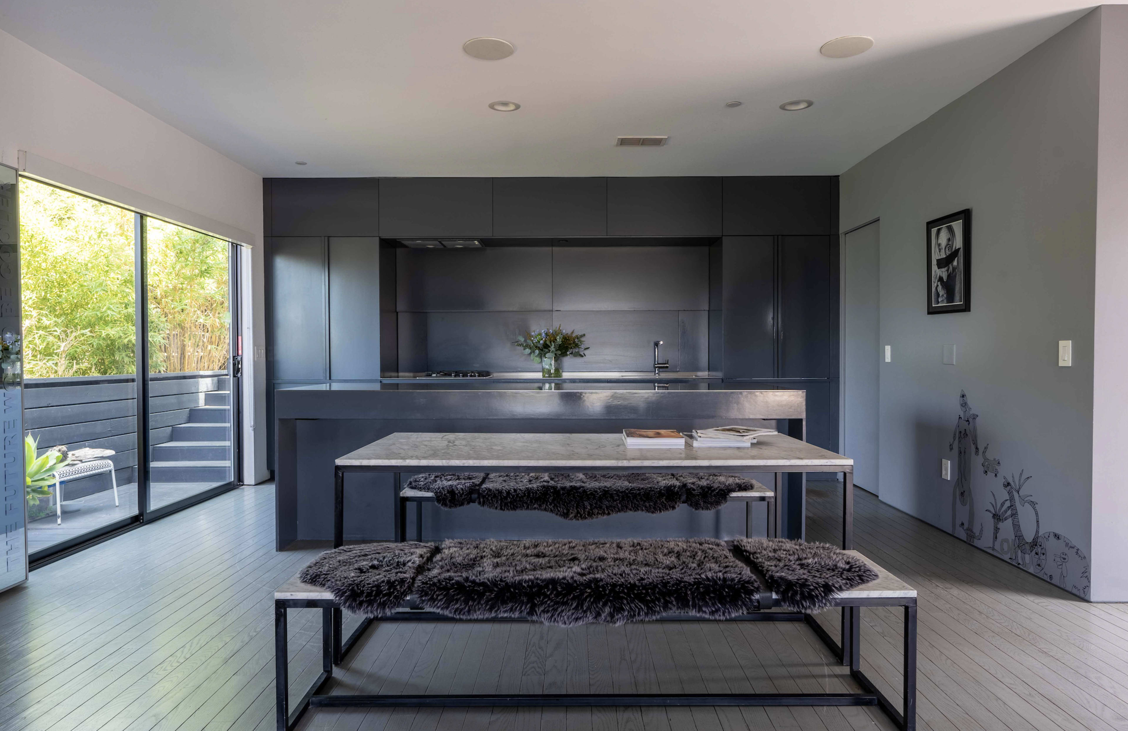 A modern kitchen features a sleek black cabinetry design, a central dining table with a gray surface, and a spacious layout with large windows.