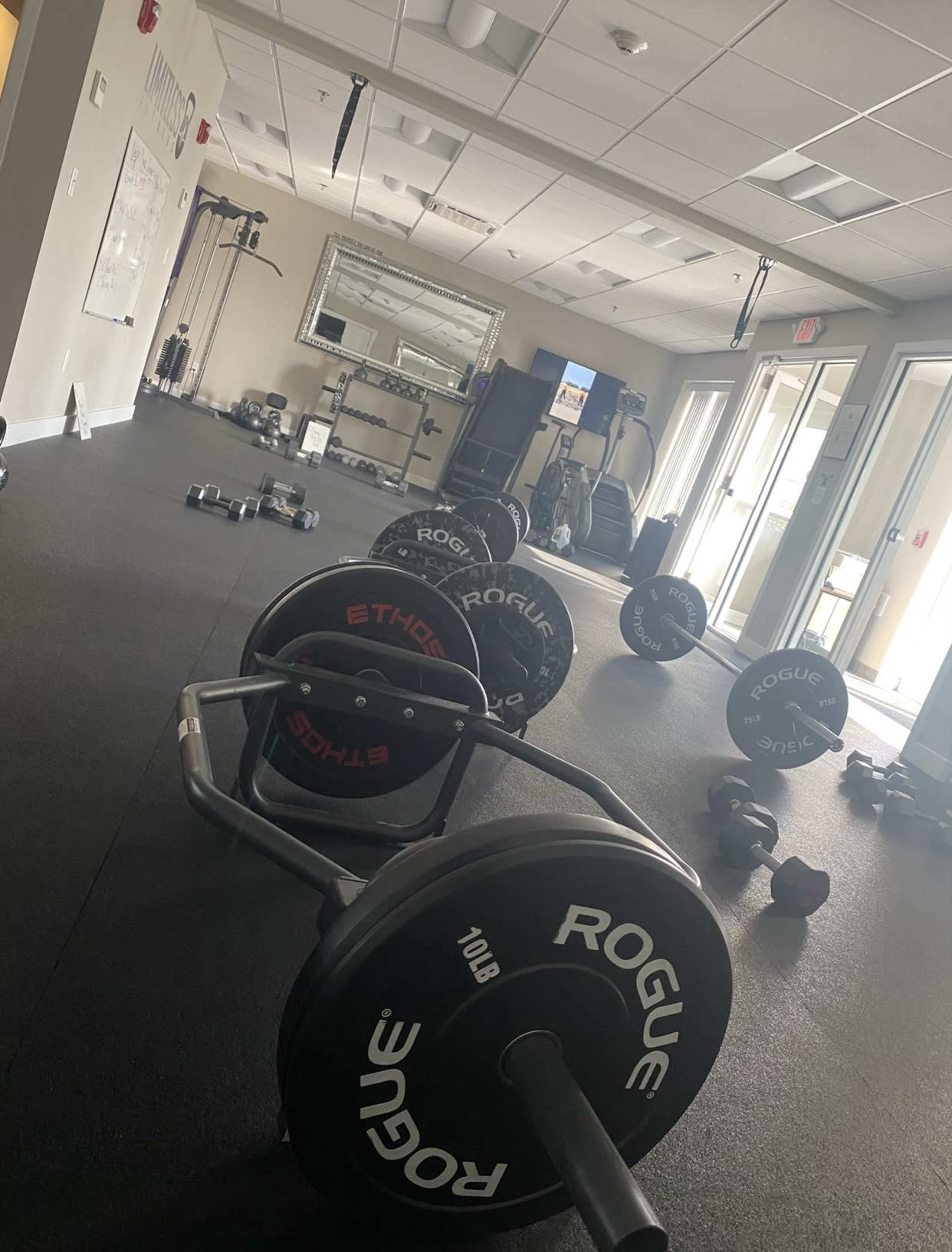 The image shows a fitness room with various weights, including barbells and dumbbells, arranged on a rubber floor, with workout equipment positioned in the background.
