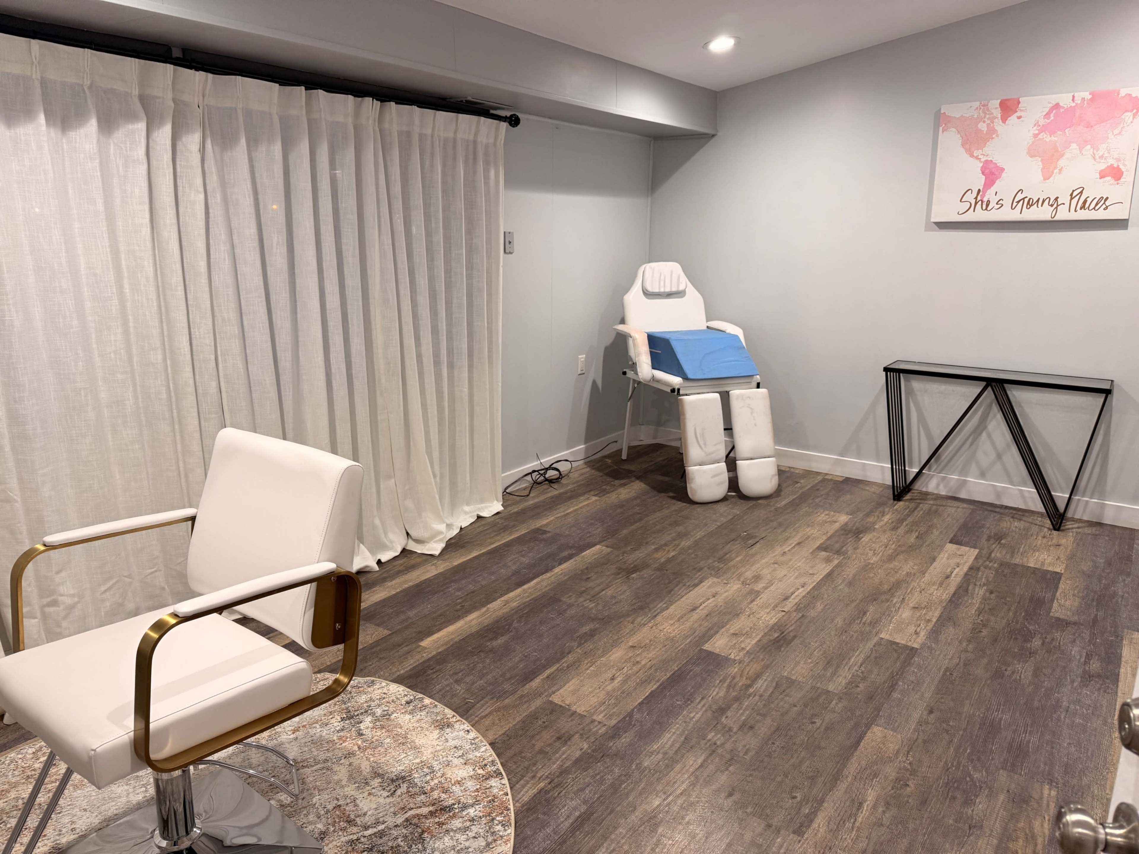 The image shows a minimalistic treatment room with a reclining chair, a white armchair, a small black table, and sheer curtains.