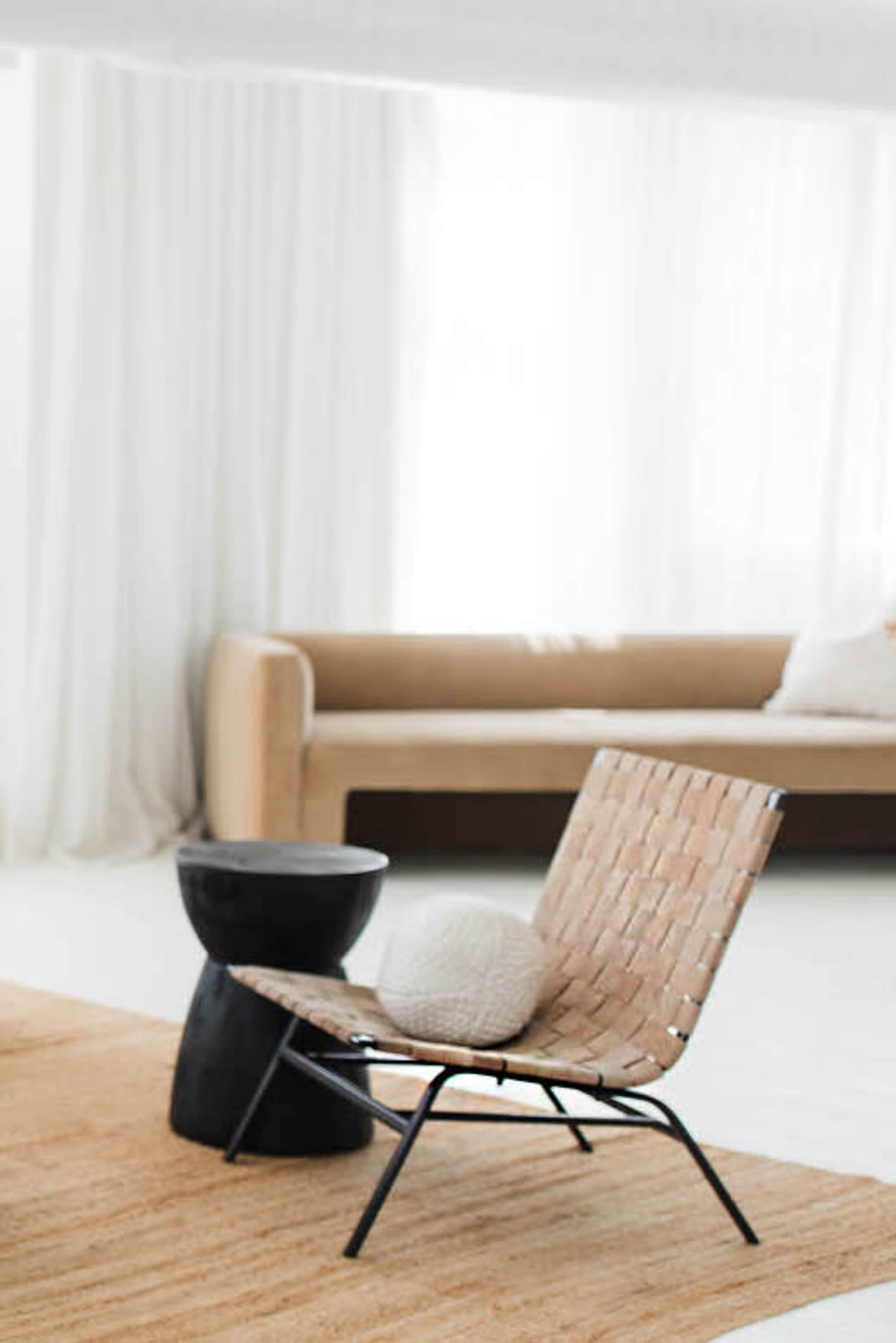 A woven chair is positioned next to a black side table on a light-colored rug in a bright, airy room with sheer white curtains.