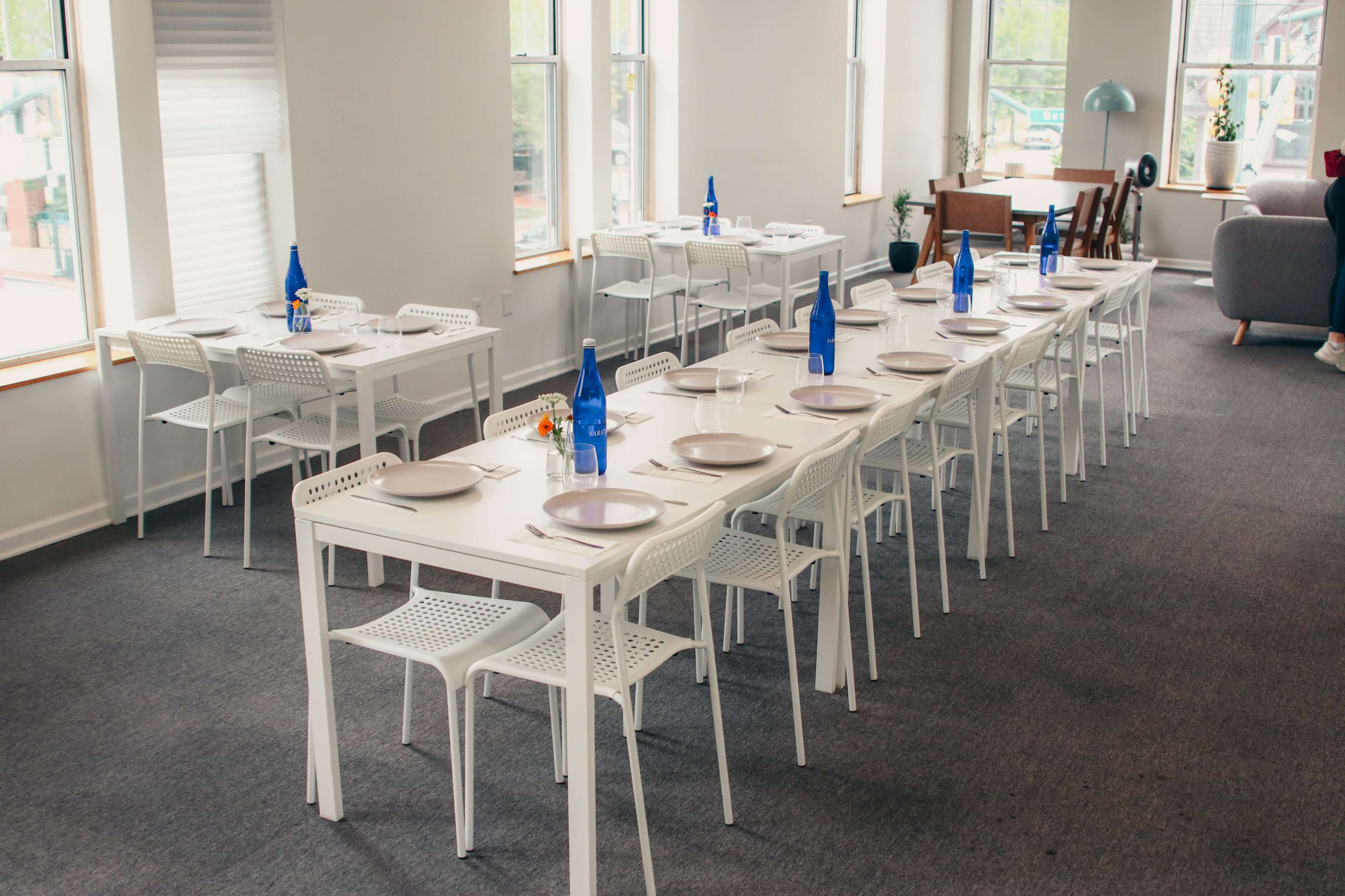 A series of white tables set up in a bright room with blue glass bottles and plates arranged neatly on each table.