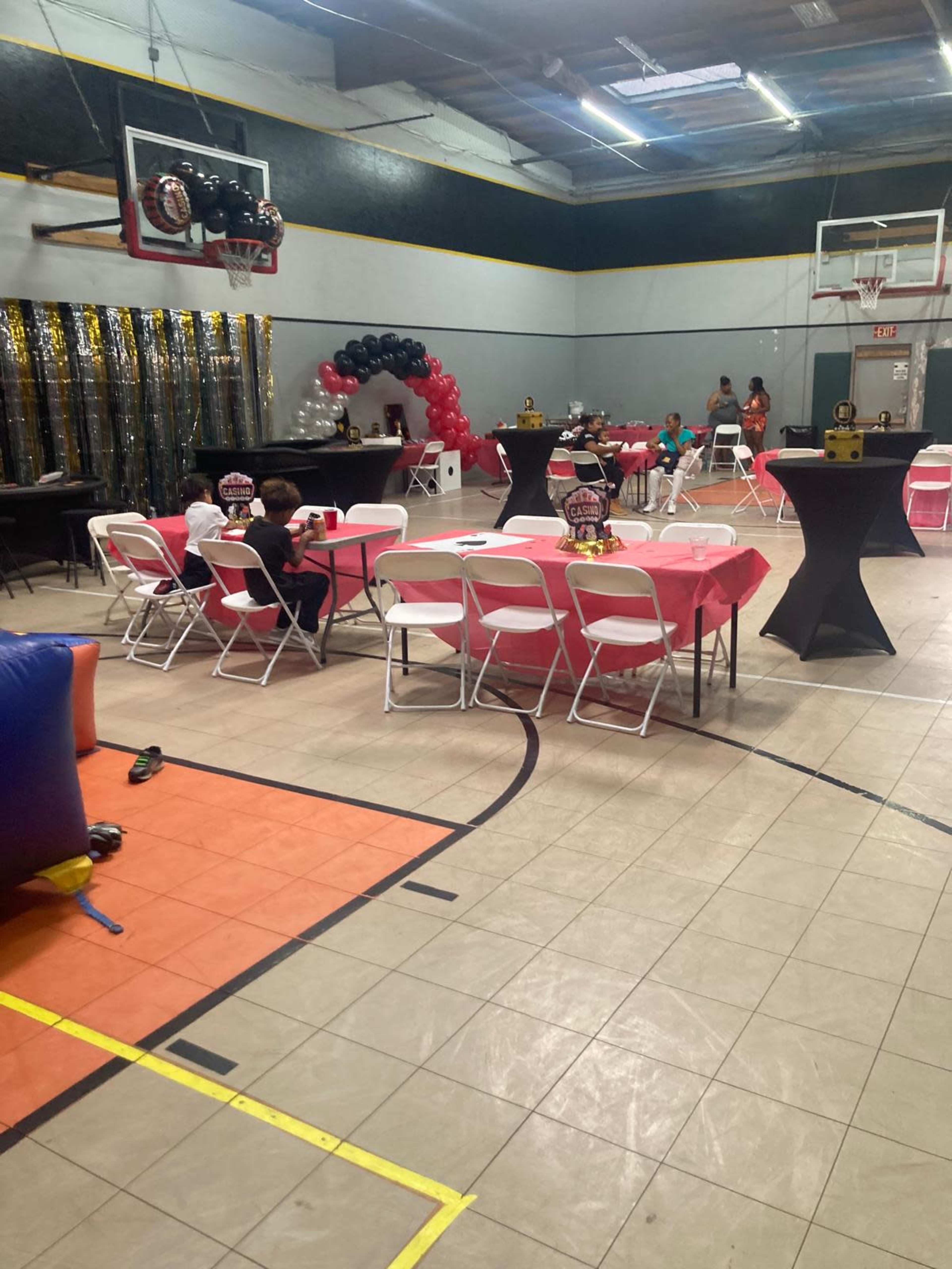 The image shows a decorated gymnasium with tables covered in red tablecloths, black and red balloons, and children seated near the tables, while others are engaged in activities in the background.