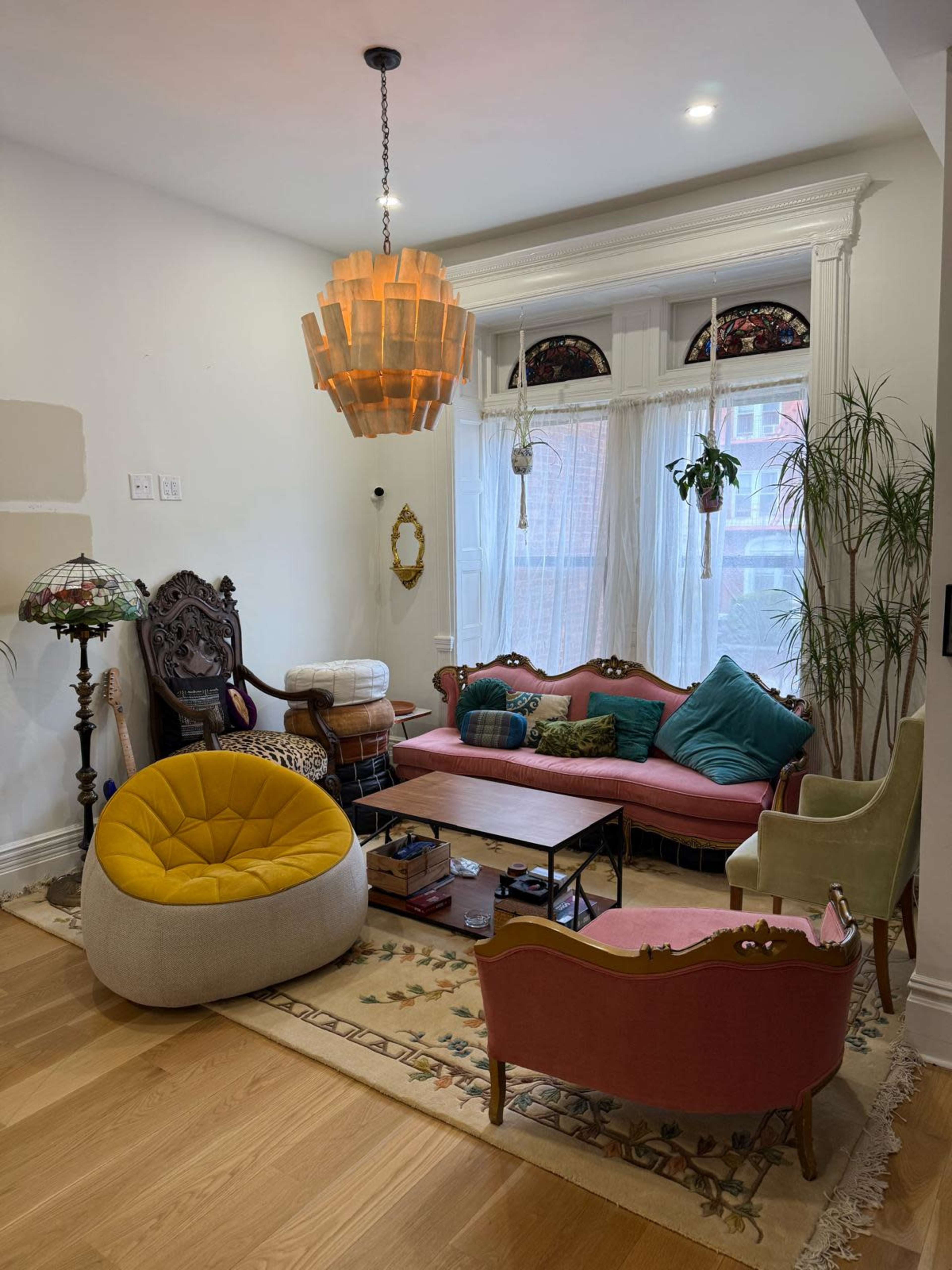 The image shows a cozy living room with a mix of vintage furniture, including a pink sofa, a yellow bean bag, a wooden coffee table, and a decorative lamp hanging from the ceiling.