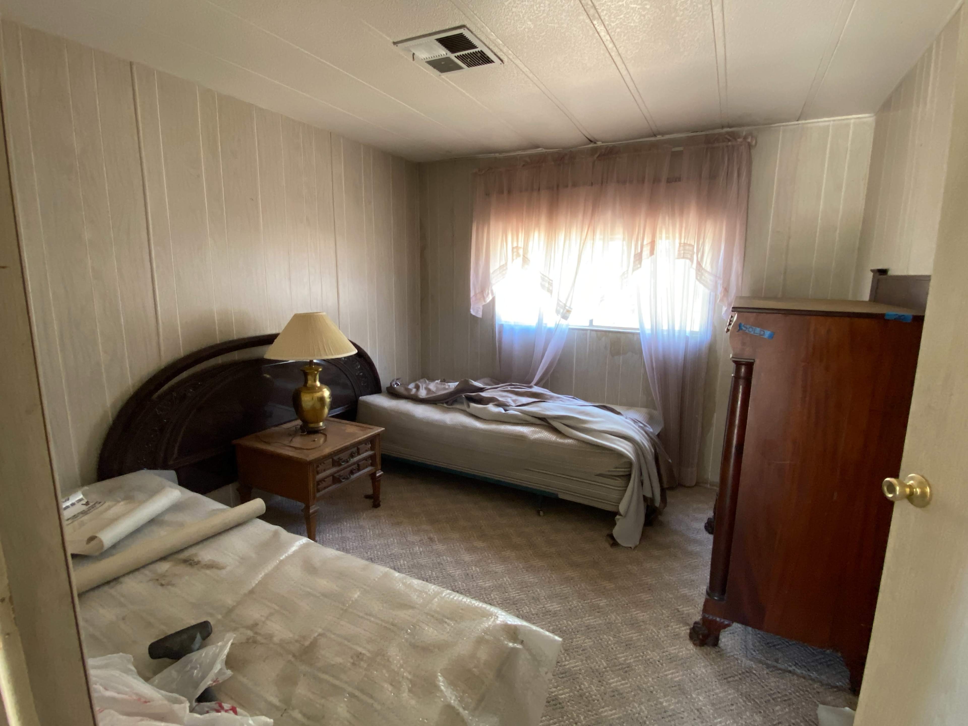 The image shows a sparsely furnished bedroom with a large window, two beds, a wooden dresser, and a lamp on a nightstand.