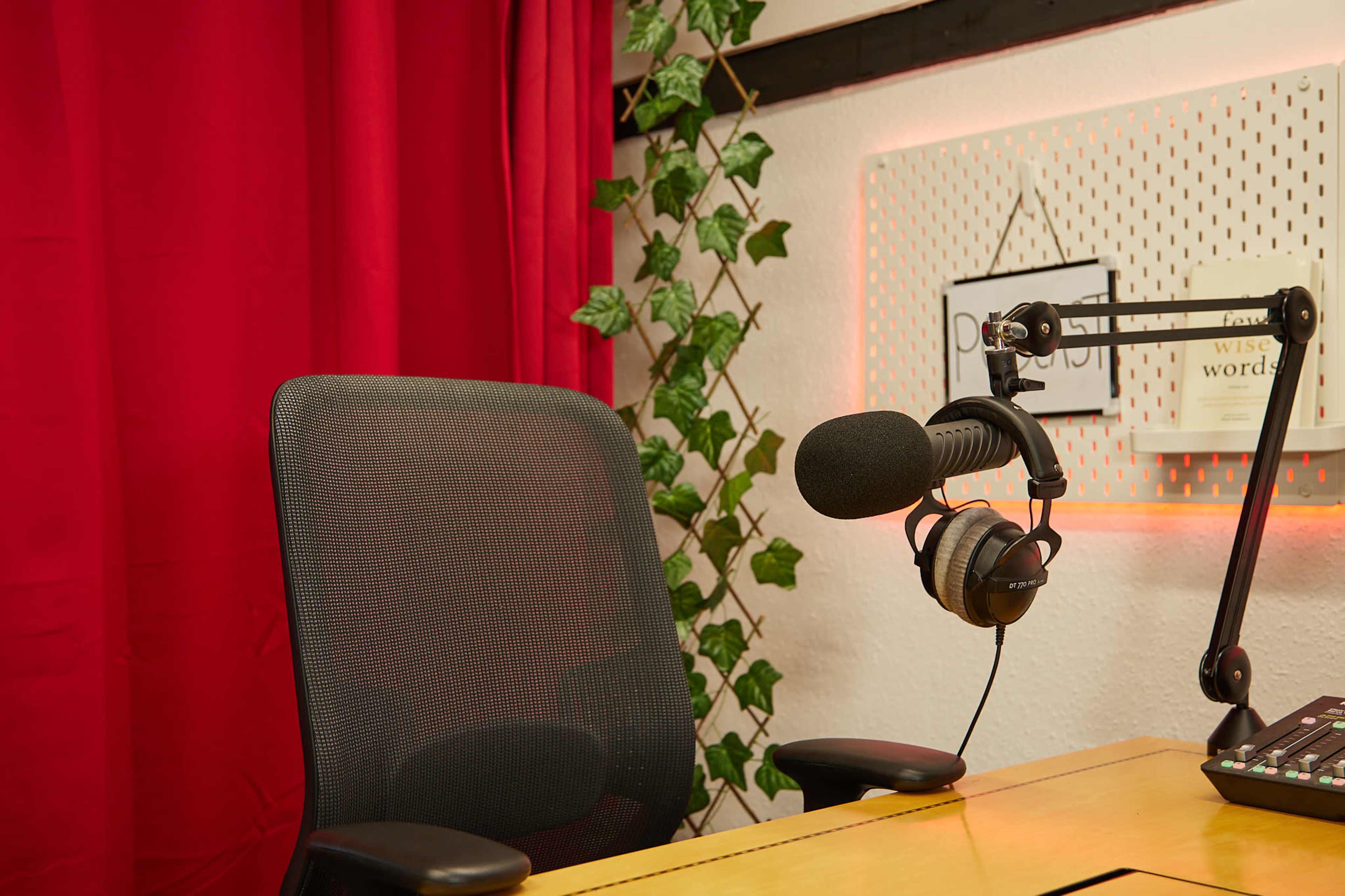 The image shows a microphone mounted on an adjustable arm next to an empty office chair in a room with red curtains and a wall adorned with a plant and books.