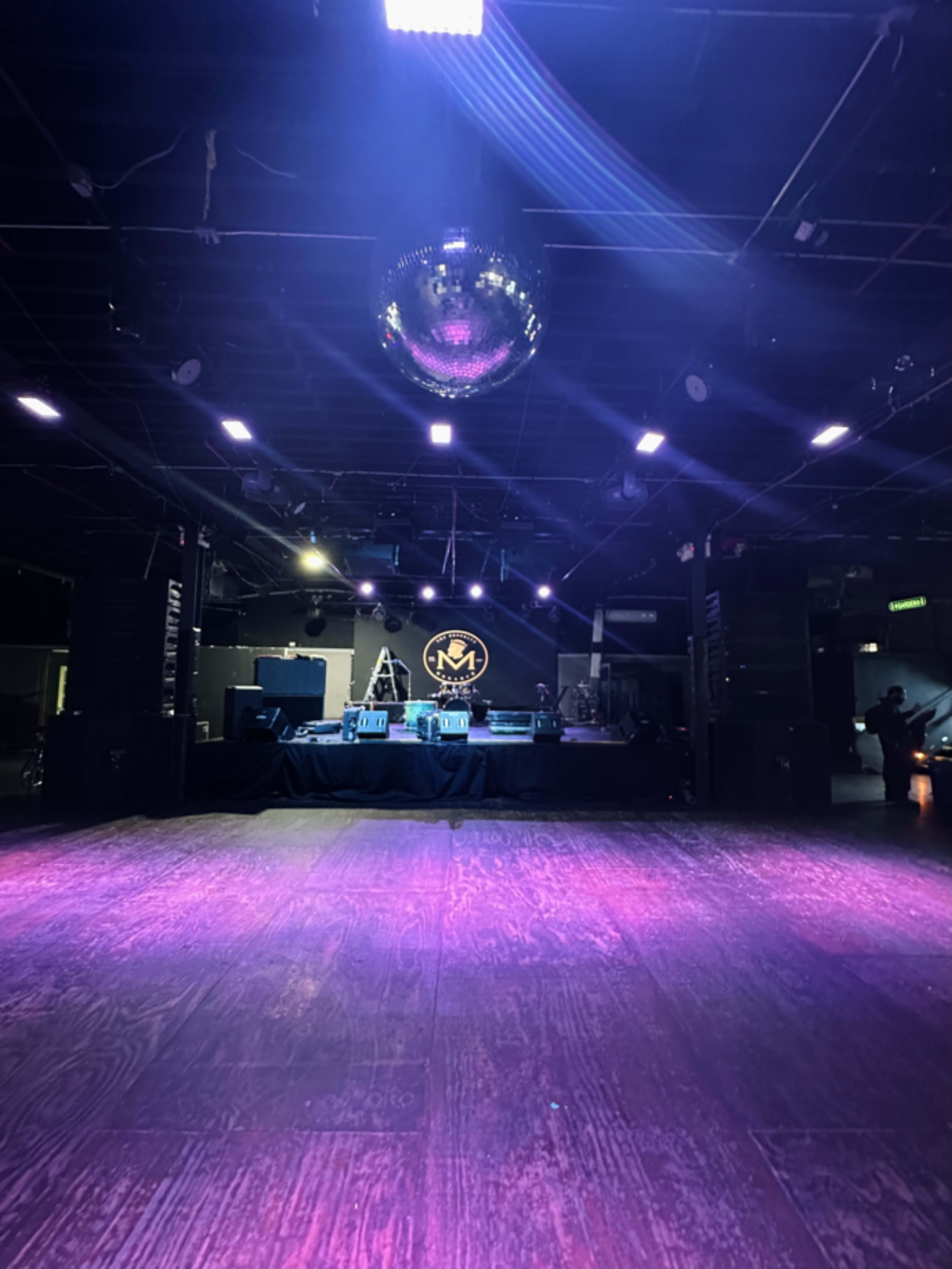 An empty stage area featuring a disco ball, illuminated by purple and white lights, with a sound system and equipment set up in the background.